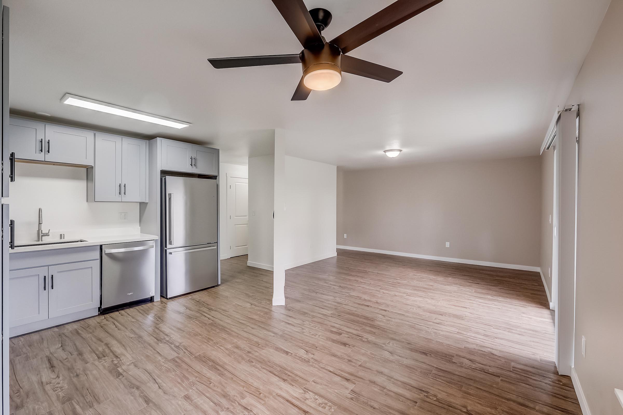 Modern interior of an apartment featuring a kitchen with light gray cabinets and stainless steel appliances. The open living area has wood-like flooring and a ceiling fan, with neutral walls and ample natural light. A door leads to a patio or balcony area.