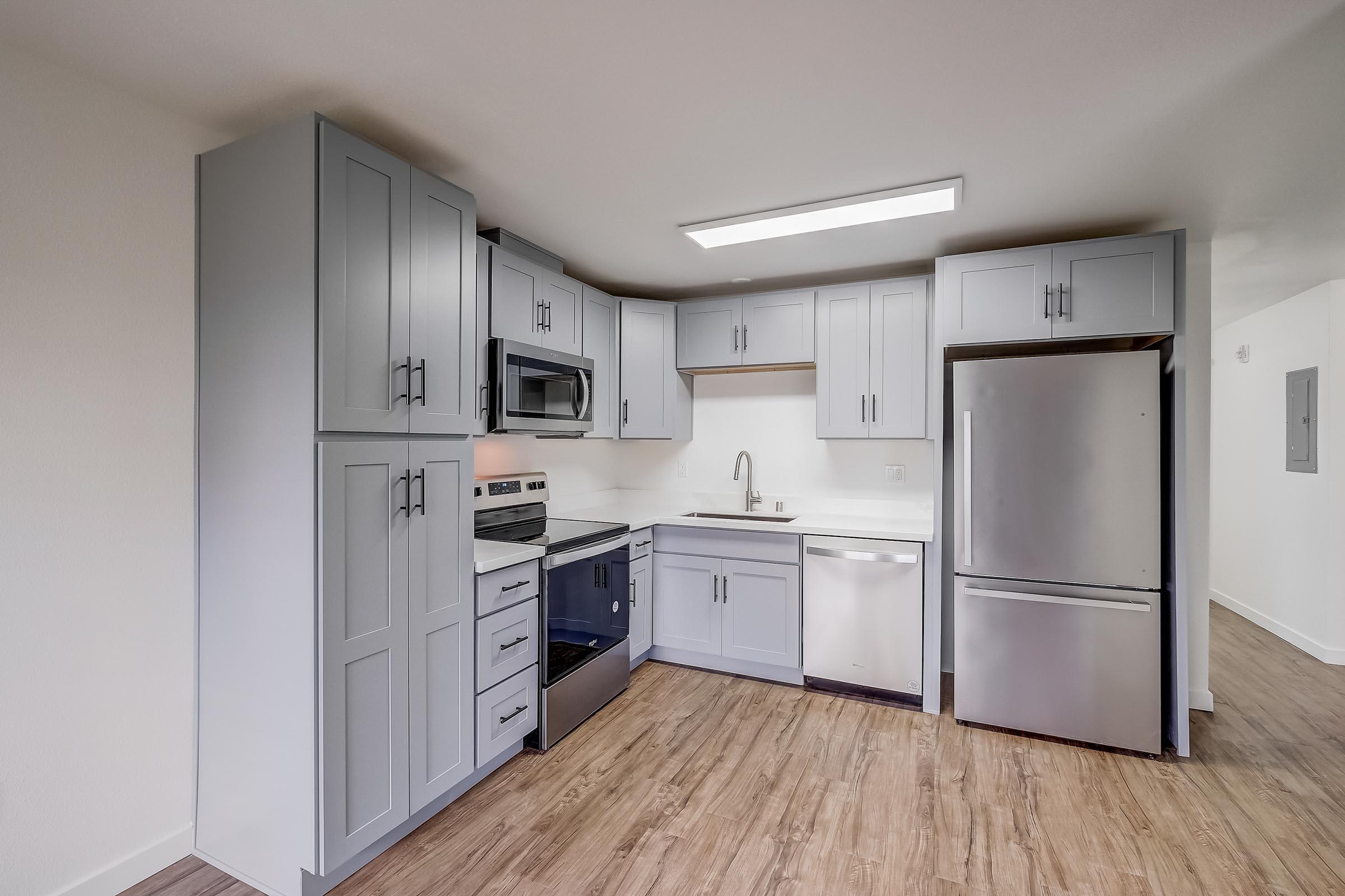 Modern kitchen featuring light gray cabinets, stainless steel appliances including a microwave, oven, refrigerator, and dishwasher. The countertop is white, and the floor has a wood-like finish. The design is spacious and contemporary, with good lighting from a ceiling fixture.