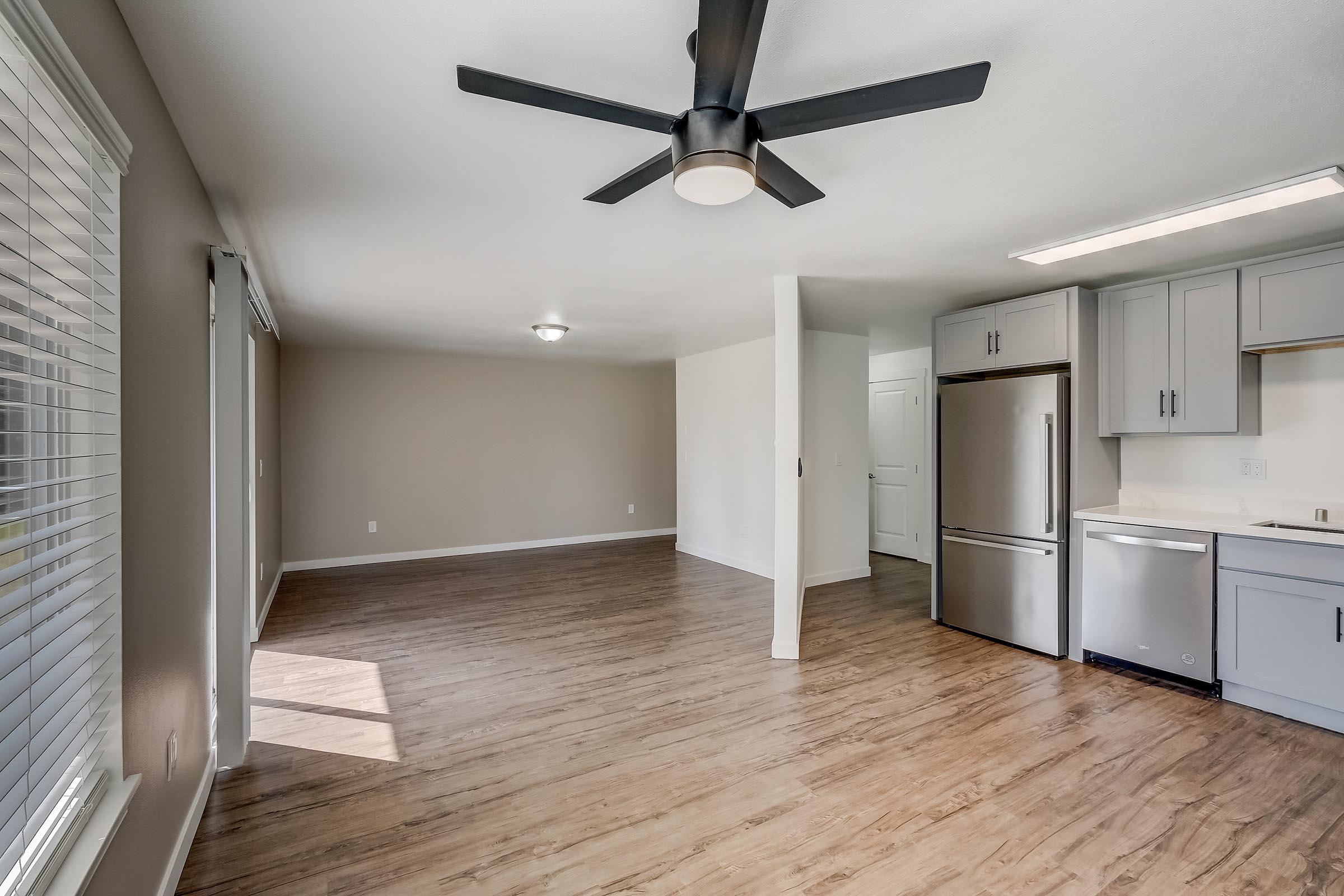 Spacious interior of a modern apartment with light-colored walls and wood-style flooring. The room features a ceiling fan, large windows with blinds, and an open kitchen area that includes stainless steel appliances and cabinetry. Natural light fills the space, creating a warm atmosphere.