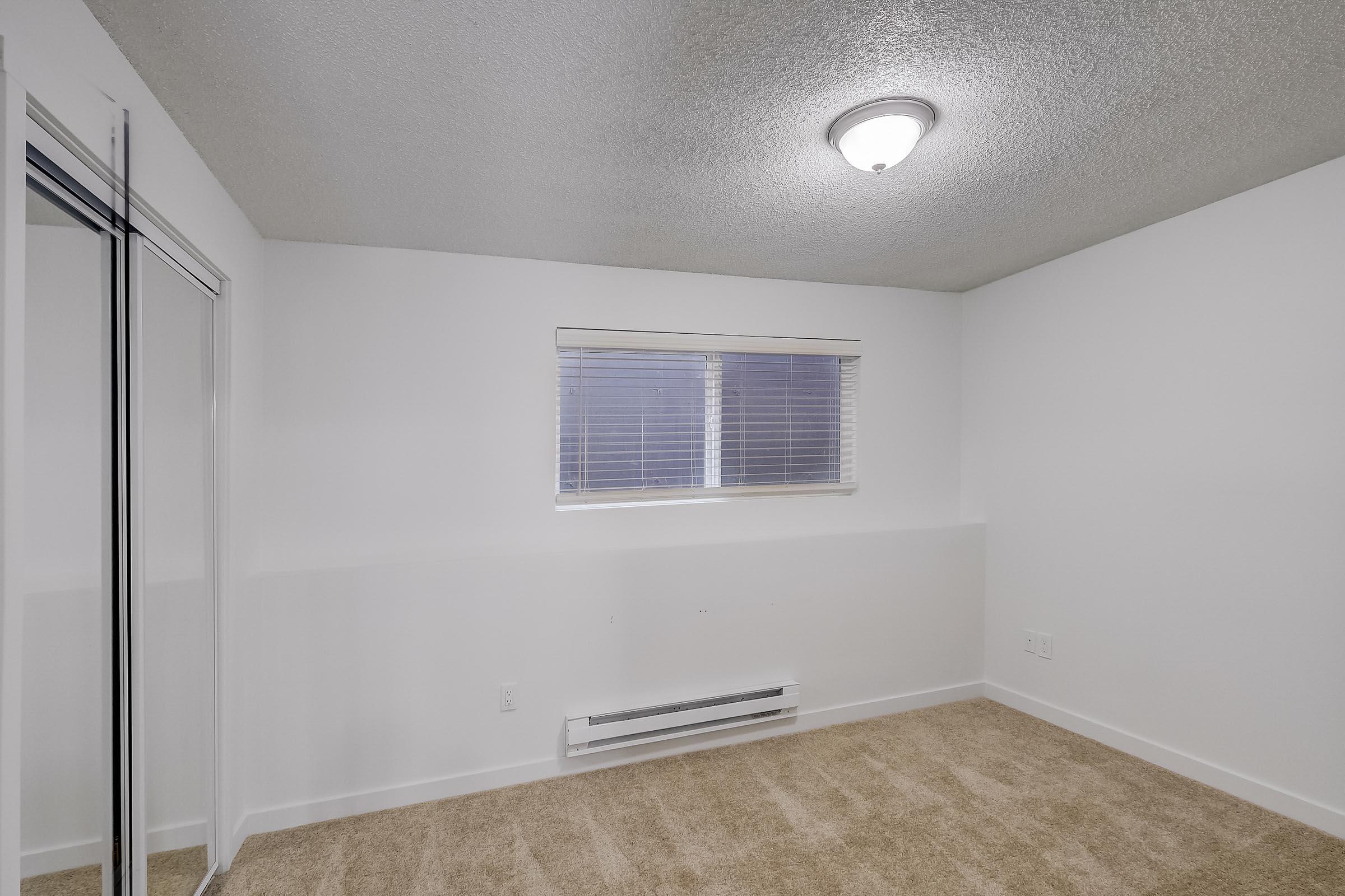 A small, empty room featuring light beige carpet, a window with blinds, and a ceiling light fixture. There is a closet with sliding glass doors on the left side. The walls are painted white, and the room appears well-lit and clean, offering a simple and modern aesthetic.