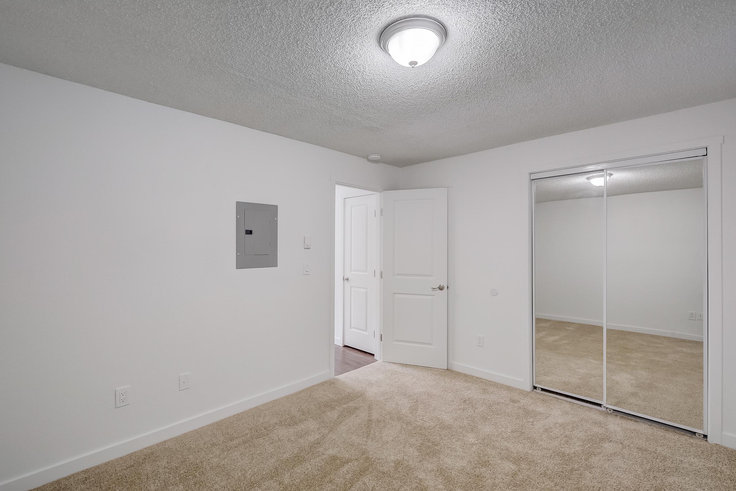 A small room featuring a light-colored carpet and white walls. There's a ceiling light fixture, a mirrored closet with sliding doors, and an electrical panel on the wall. A door leads to another space, and the overall ambiance is clean and minimalistic.