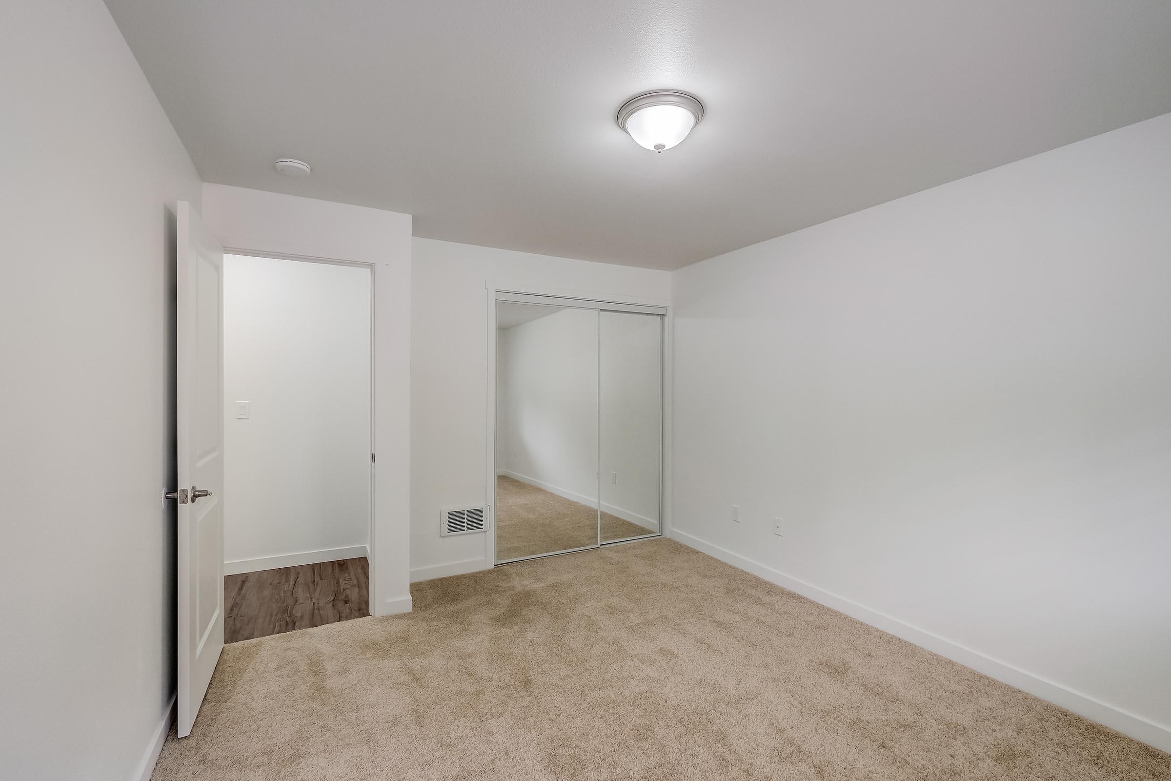 A spacious, empty room featuring light beige carpet, white walls, and a ceiling light fixture. A doorway leads to a small adjacent room, while a wall with mirrored sliding doors enhances the sense of space. The overall design is simple and modern, perfect for personalization.
