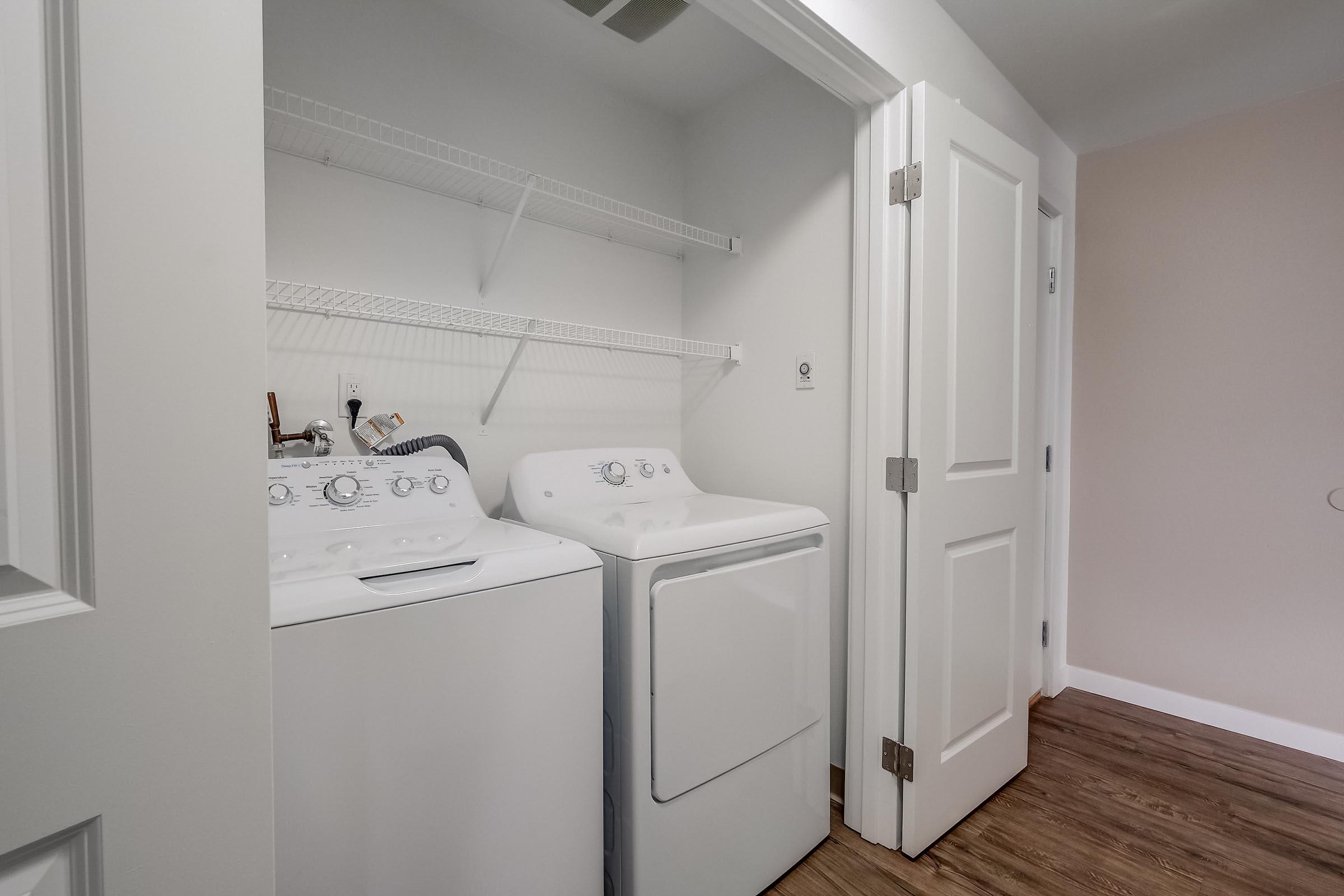 A laundry room featuring a washer and dryer side by side, with a wire shelf above for storage. The room has light-colored walls and a wooden floor, with two doors visible, one leading to the main area and the other potentially a closet or utility space.