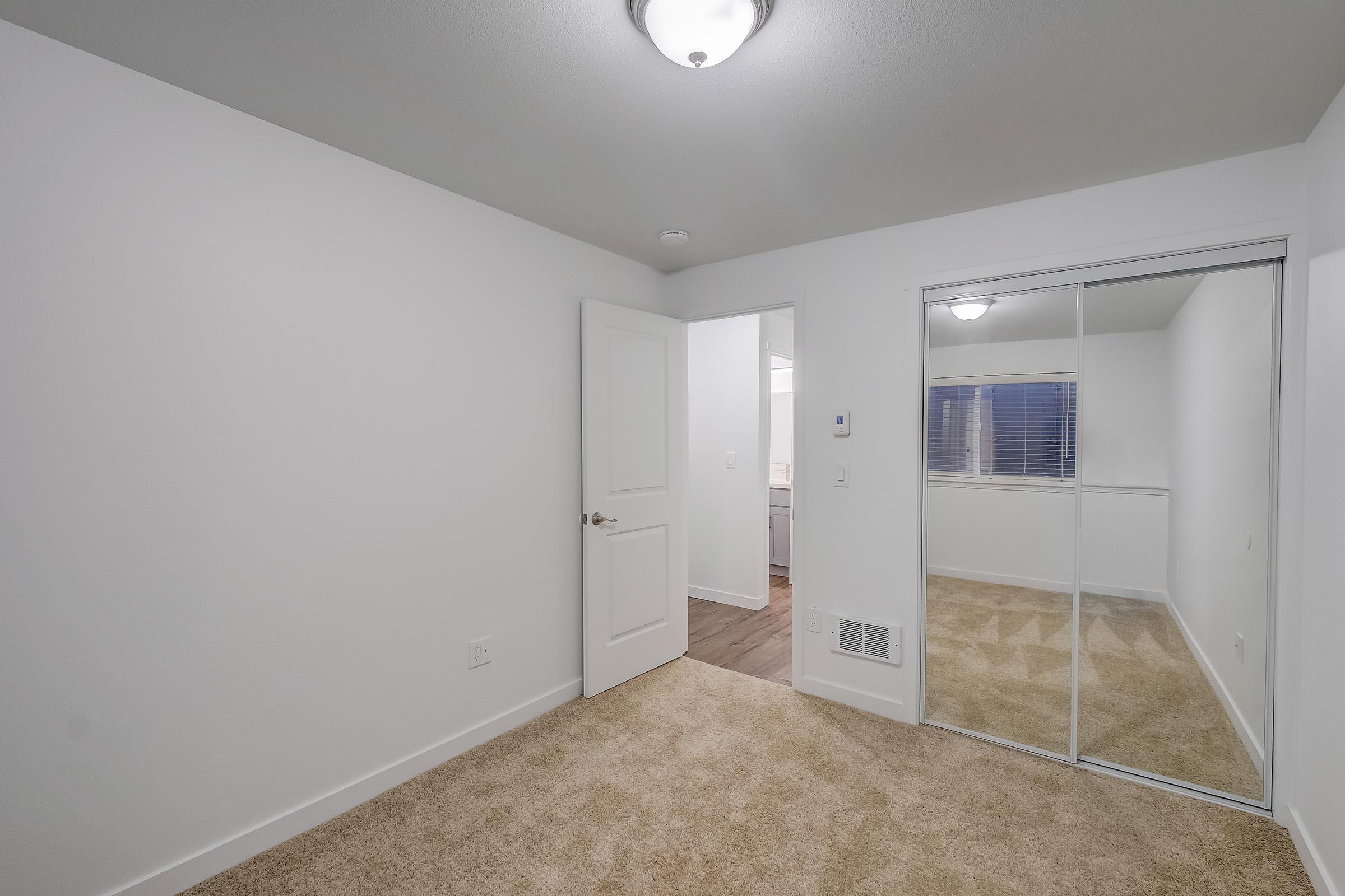 A bright, empty room with light-colored walls and plush beige carpet. There is a closet with sliding mirrored doors on one side, and a partially open door leading to another room or bathroom. A ceiling light fixture is visible, providing illumination. Natural light filters in through a window.