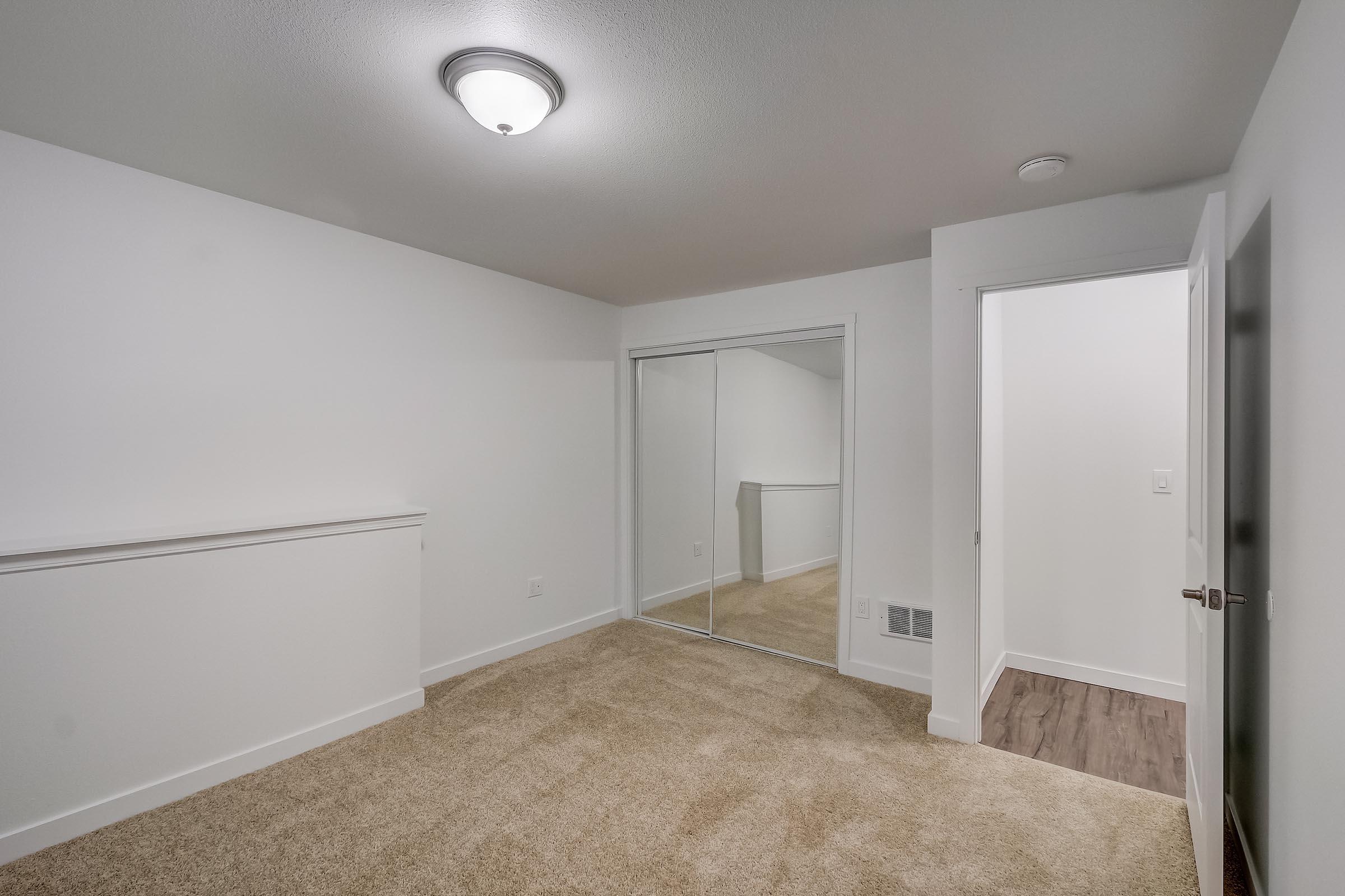Empty interior of a room with light-colored walls and carpet flooring. Features a ceiling light fixture, a mirrored sliding closet door, and a doorway leading to another room. The space is uncluttered and well-lit, suggesting a modern design.