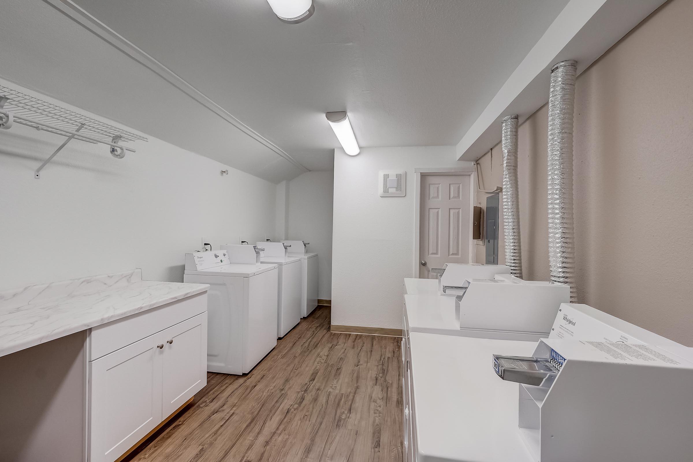A clean and organized laundry room featuring several white washing machines and dryers. The space includes a countertop, wall-mounted shelves, and a door leading to another area. The floor is wooden-style, and there are aluminum vents on the wall. Bright overhead lighting enhances the bright, airy atmosphere.