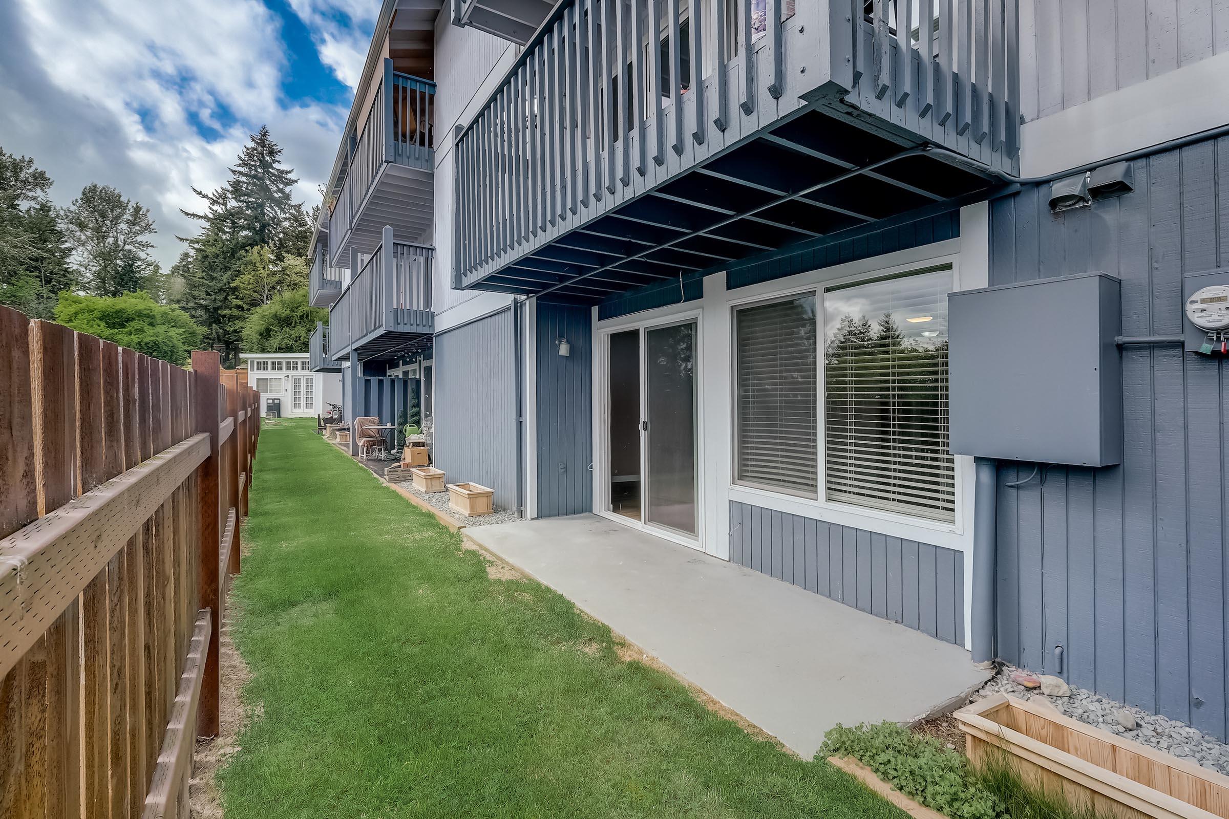 View of a modern residential exterior featuring a patio area with sliding glass doors, a well-maintained lawn, and wooden fencing. The building showcases multiple balconies and a serene atmosphere with trees in the background under a partly cloudy sky.