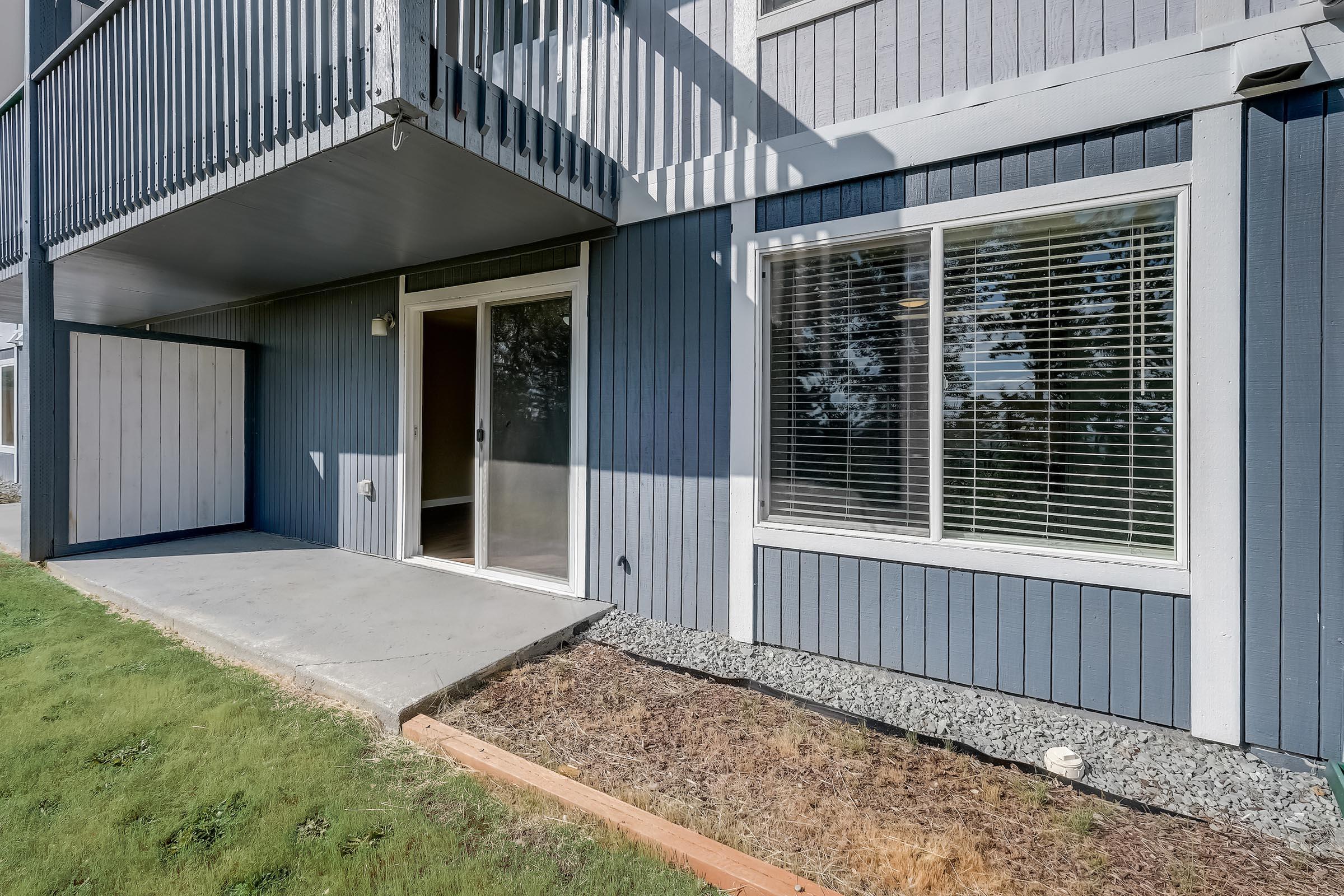 Ground-level view of a modern building exterior featuring a patio door and large windows with white blinds. The wall is painted in blue tones, and there is a small grass area in front of the building, along with a gravel border. The setting is bright and well-lit, indicative of a residential space.