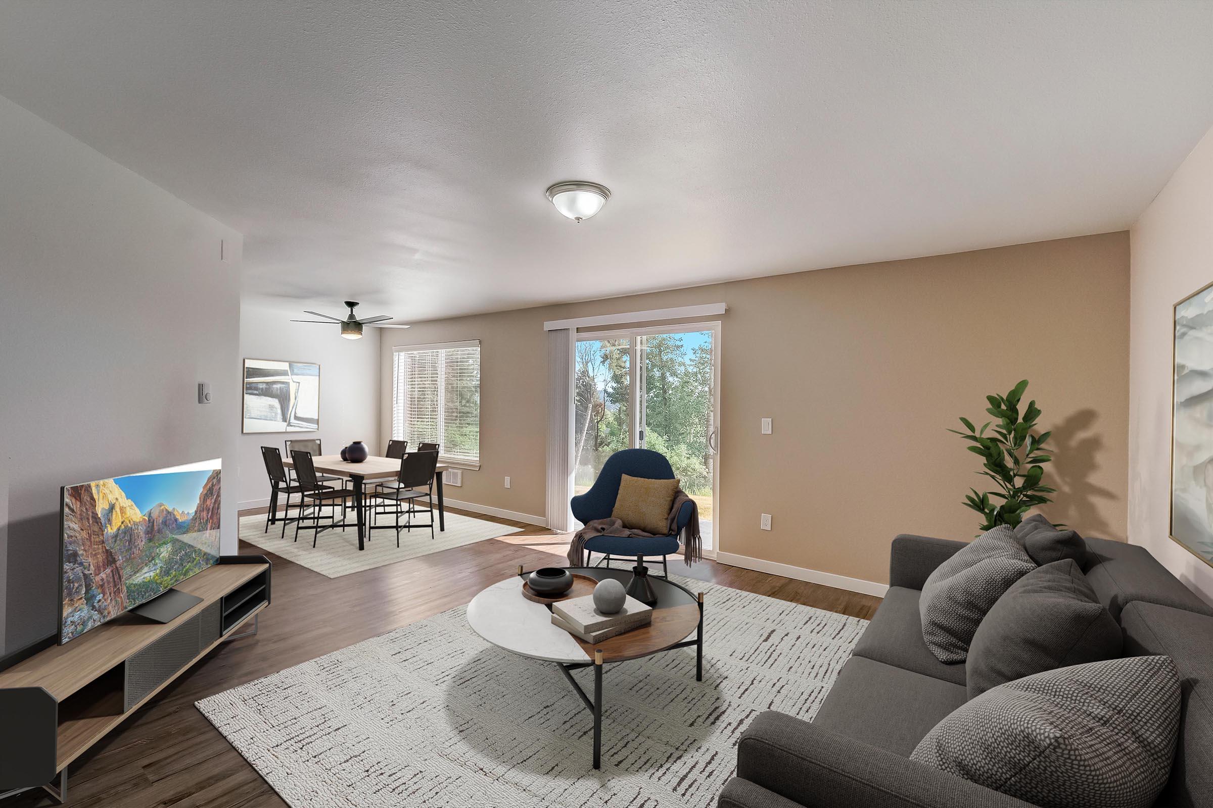 Modern living room with a gray sofa, round coffee table, and a television on a wooden stand. A dining area with a table and chairs is visible. Large windows allow natural light to fill the space, showcasing a view of greenery outside. The walls are painted a light tan color, creating a warm atmosphere.