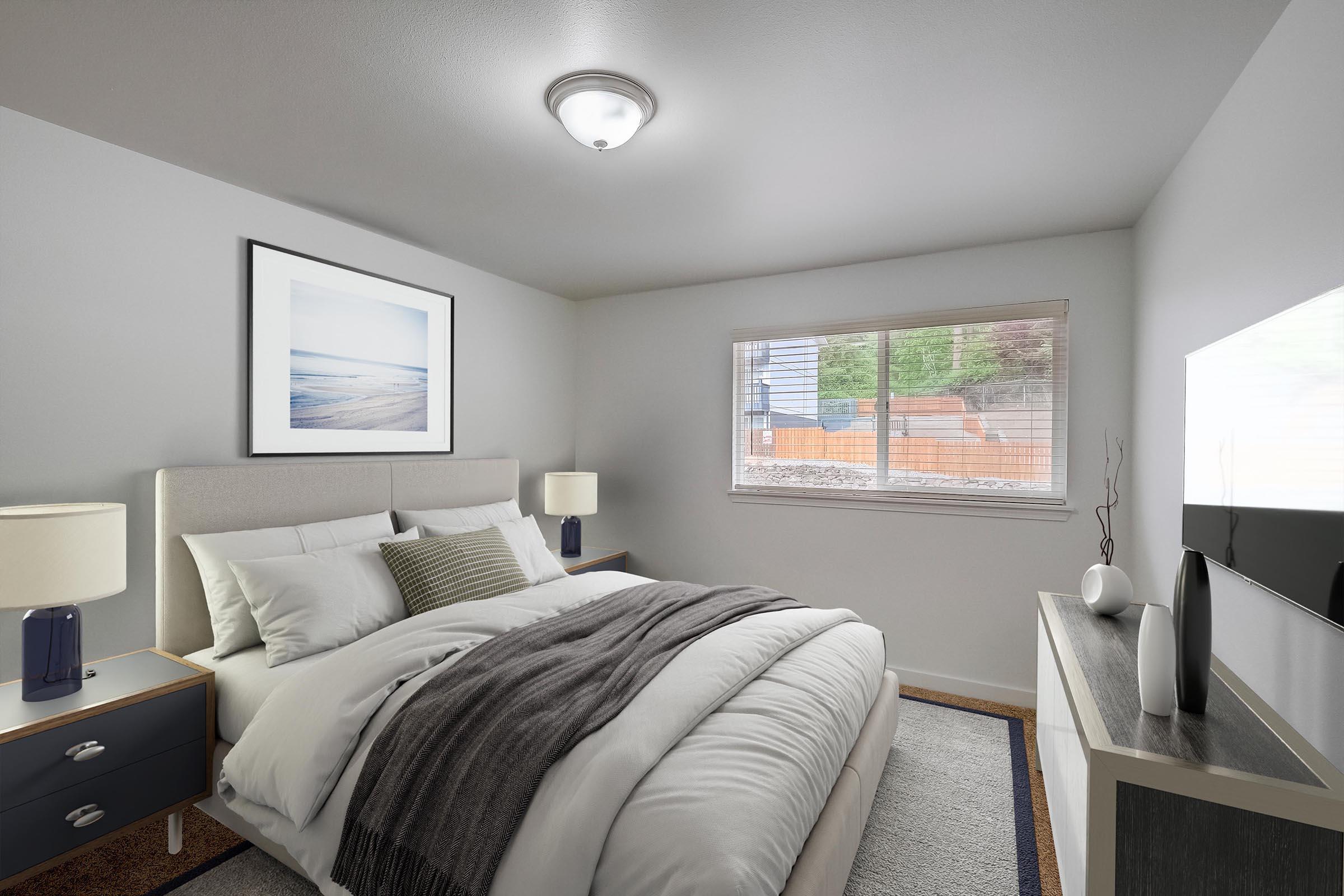 A cozy bedroom featuring a neatly made bed with white and gray bedding, a light gray headboard, and a framed ocean photograph on the wall. There are two bedside tables with blue lamps, and a modern TV stand with a television. A window allows natural light, showing greenery outside.