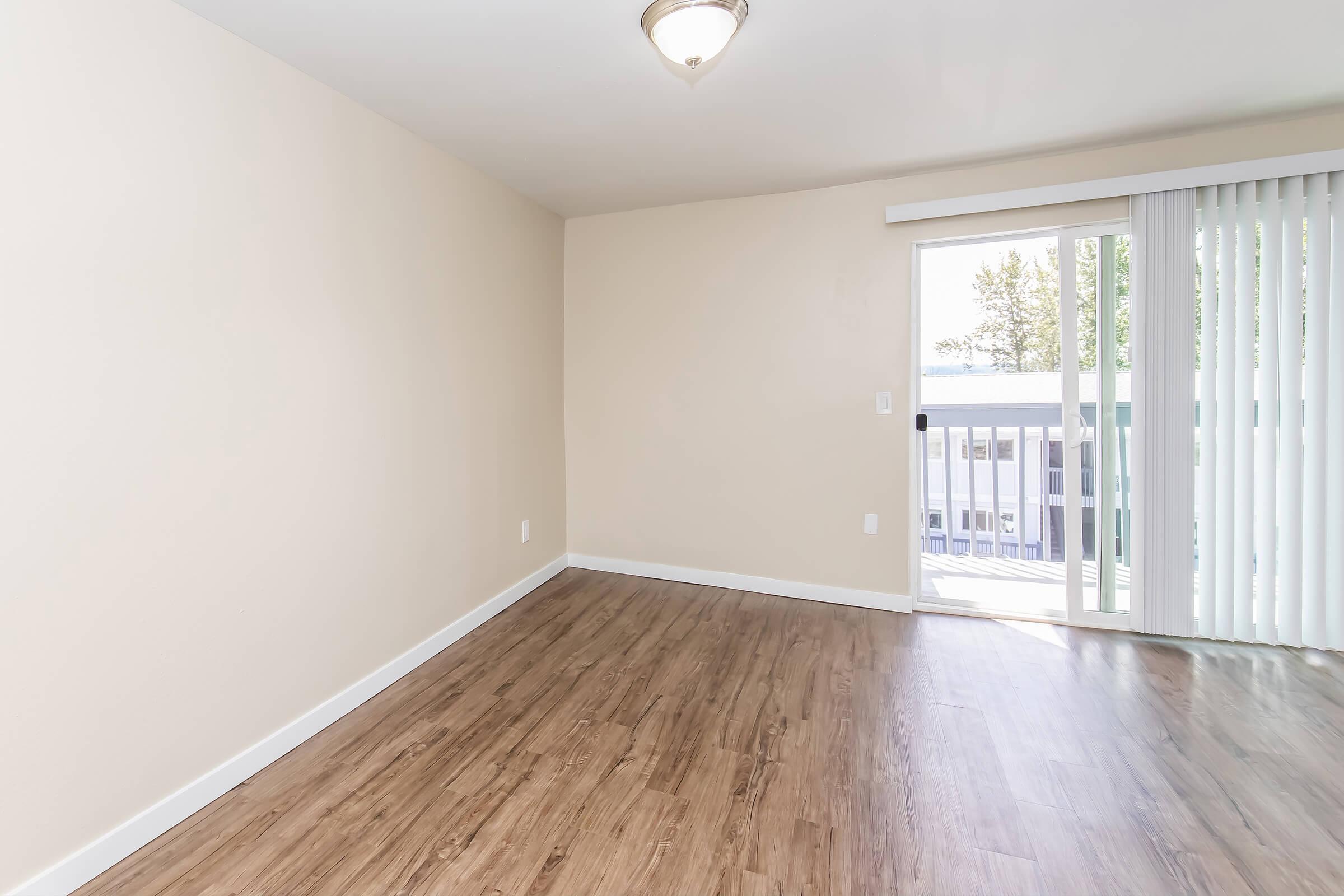 A clean, empty room featuring light beige walls and a large sliding glass door with vertical blinds. The floor is covered with light wood laminate, and a simple light fixture hangs from the ceiling. Sunlight streams in through the door, illuminating the space.