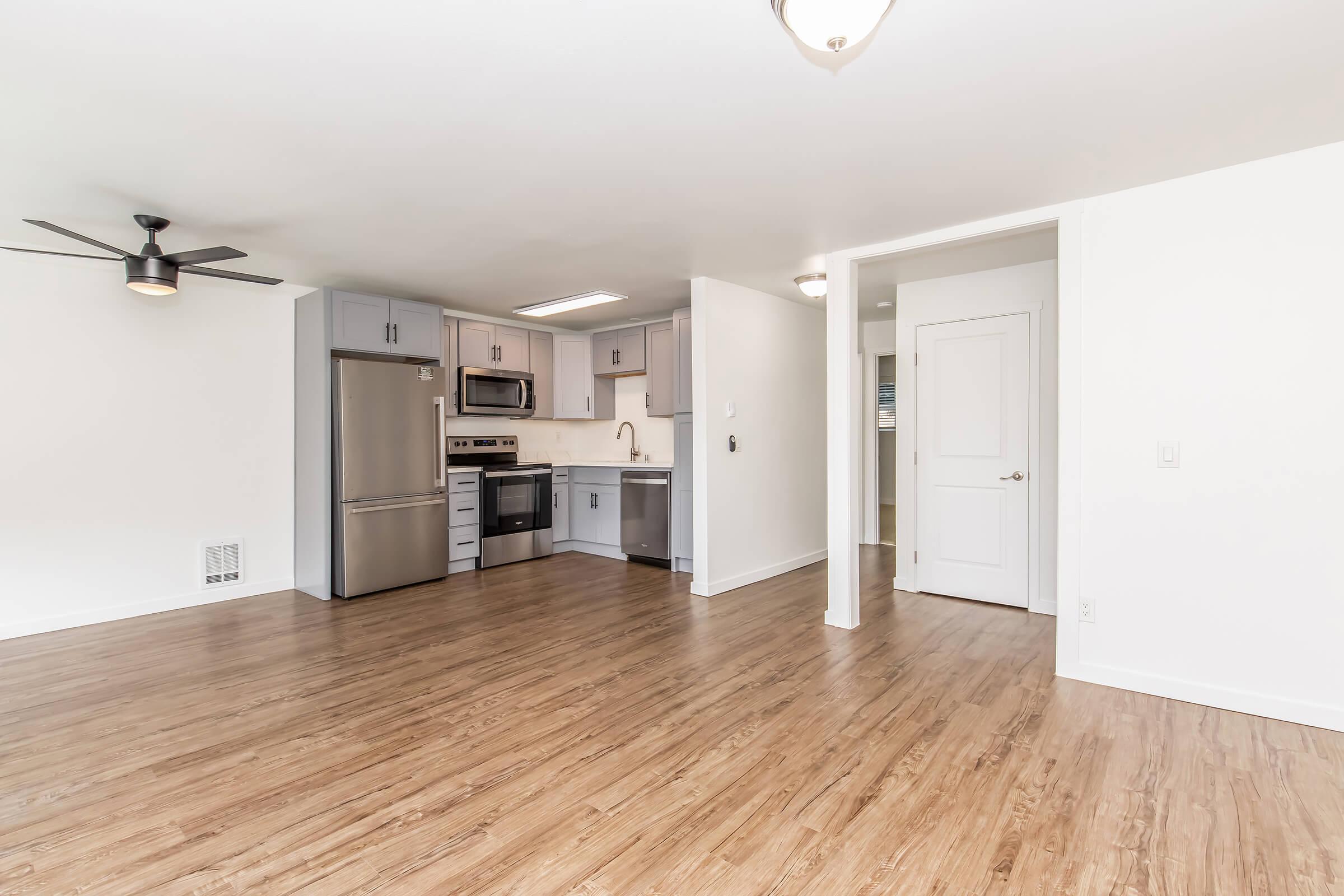 Modern kitchen area in a spacious apartment. Features stainless steel appliances, gray cabinetry, and a ceiling fan. The open layout includes hardwood flooring and a doorway leading to another room. Bright, neutral walls enhance the airy feel of the space.