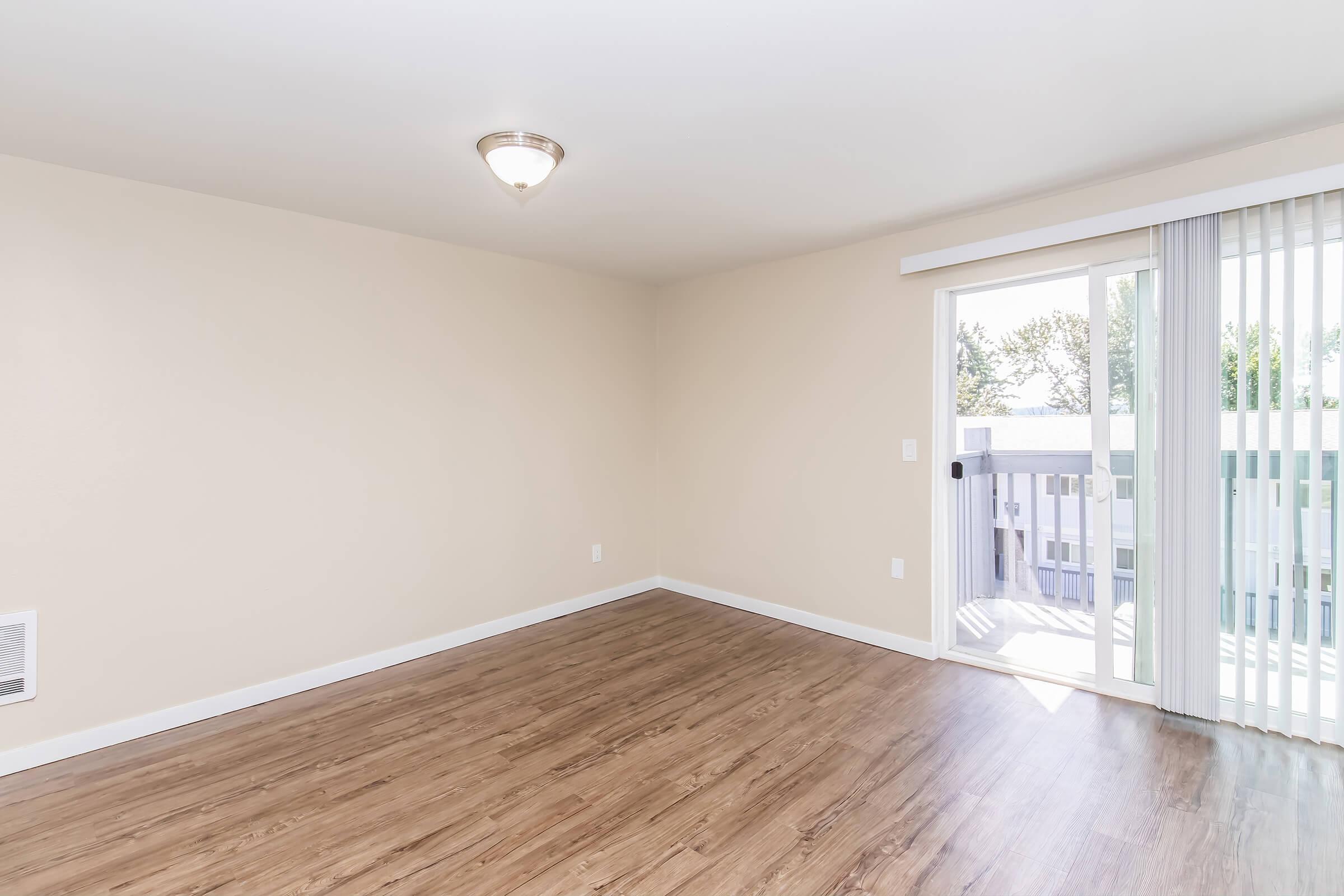 Empty room with light-colored walls and wooden laminate flooring. A ceiling light fixture is visible. There is a sliding glass door leading to a small balcony, with sheer blinds partially drawn, allowing natural light to enter the space. The overall ambiance is bright and inviting.