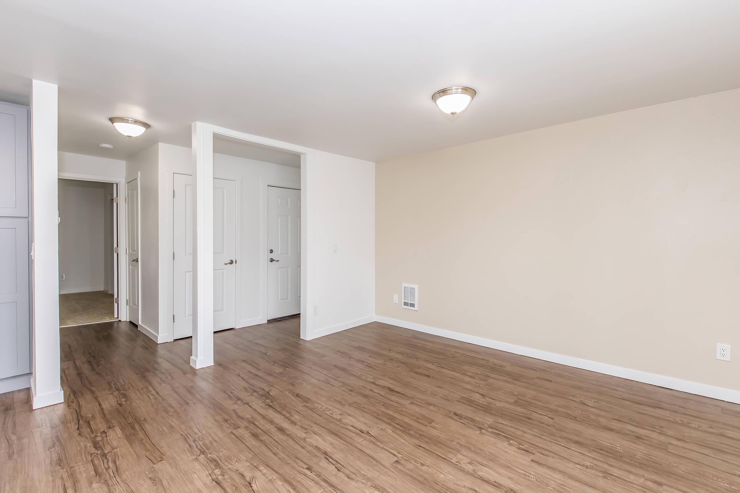 A spacious, empty room with light-colored walls and wood-style flooring. Two wall-mounted light fixtures provide illumination. On the left, there is a narrow corridor leading to a doorway. The adjacent wall is unadorned, creating a clean and inviting atmosphere.