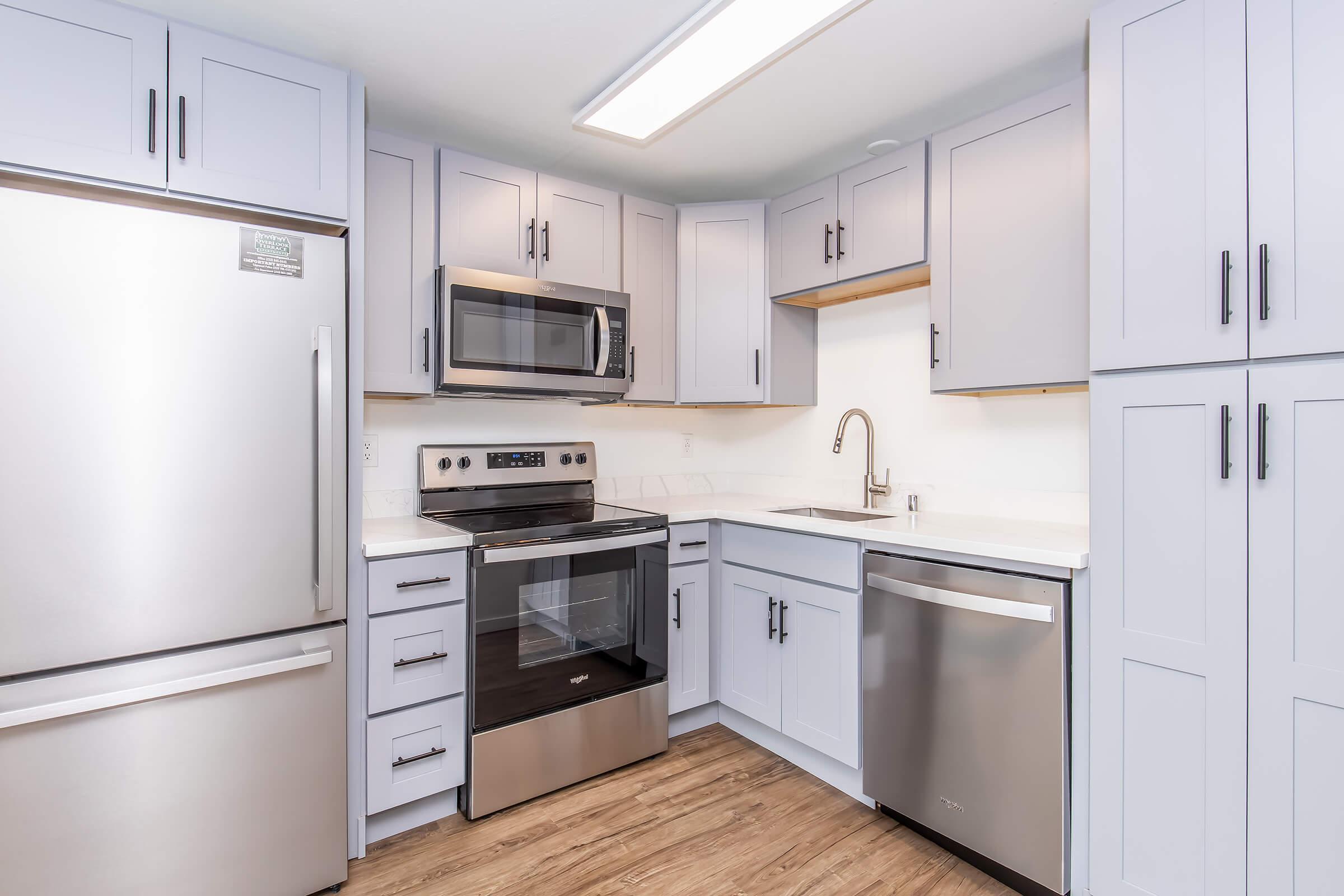 A modern kitchen featuring light gray cabinets, stainless steel appliances including a refrigerator, microwave, oven, and dishwasher. The countertops are white with a clean and contemporary design, complemented by warm wooden flooring. Natural light comes from a ceiling fixture.