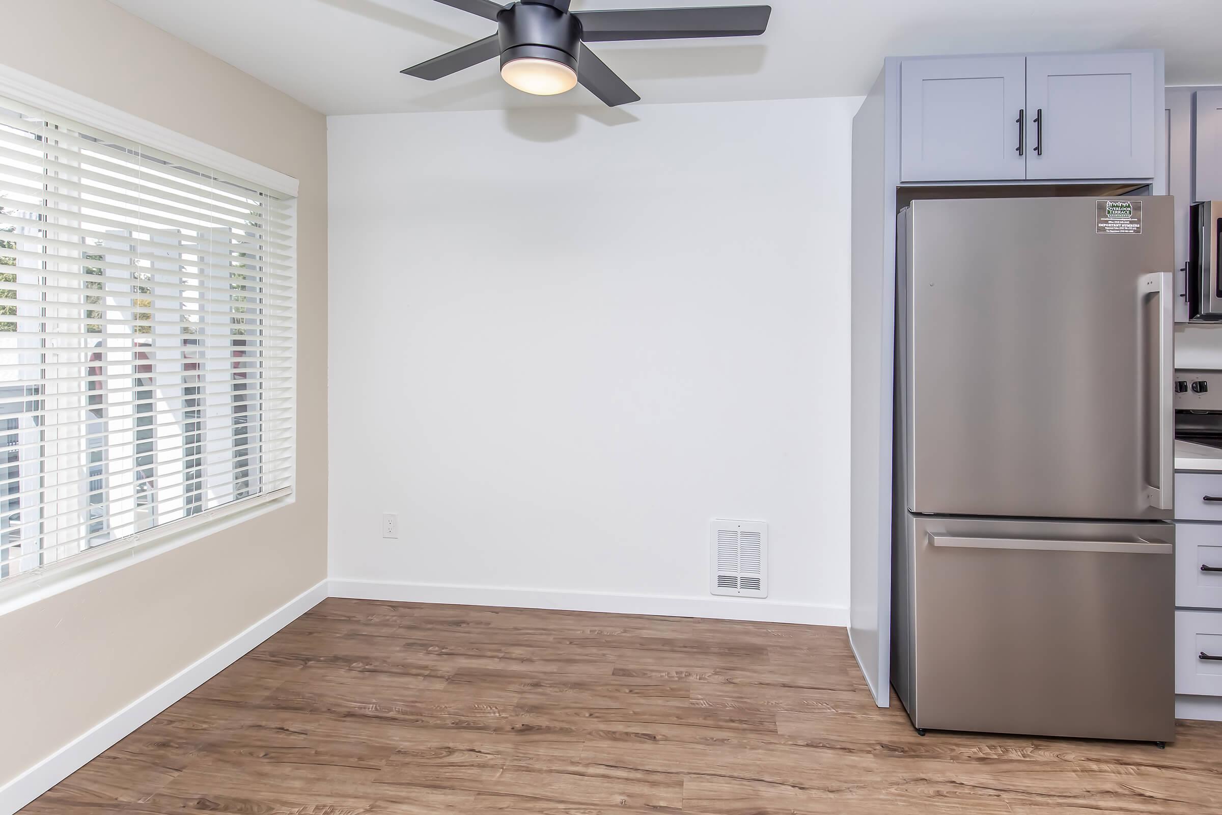 A small, modern kitchen space featuring a stainless steel refrigerator, gray cabinets, a ceiling fan with light, and large windows covered by blinds, allowing natural light to fill the room. The flooring is a warm-toned wood laminate, creating a cozy atmosphere.
