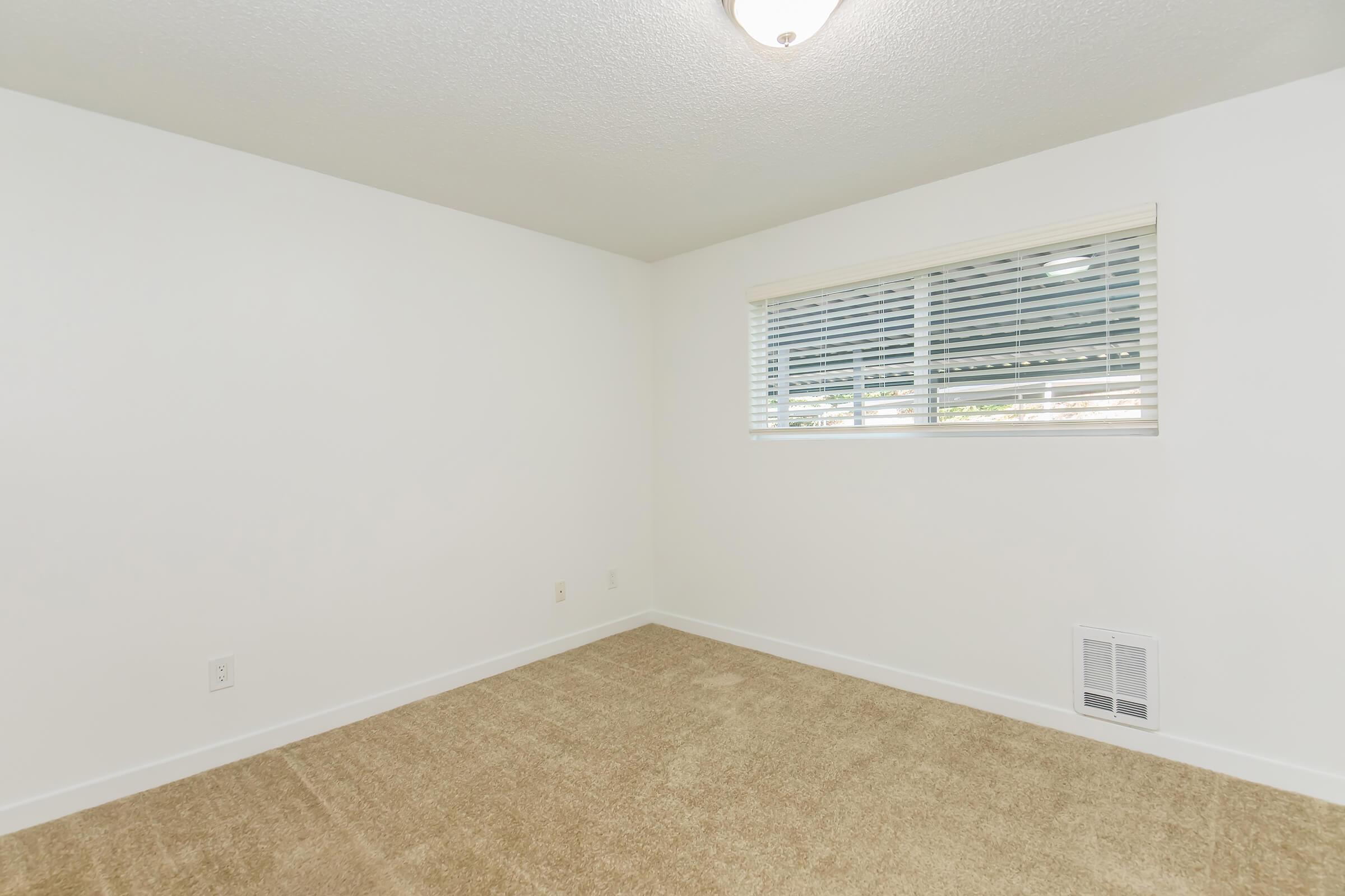 A small, empty room with light beige carpet and white walls. There is a window with horizontal blinds allowing natural light in. The ceiling has a simple light fixture, and there is a vent on the floor. The space appears clean and unadorned, suitable for various uses.