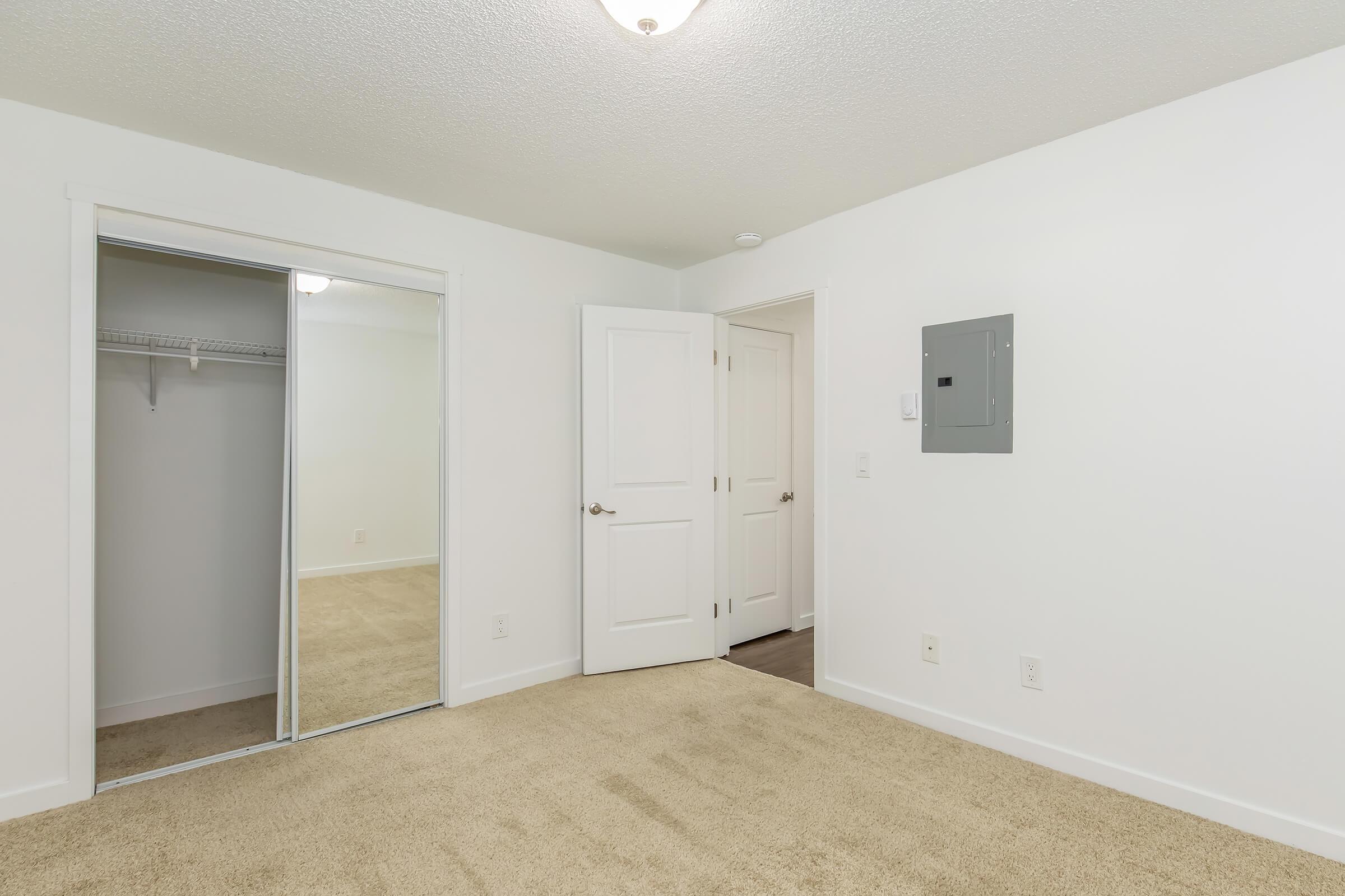 A well-lit, empty room featuring beige carpet, white walls, and a ceiling light. There are two doors: one leading to a closet with mirrored sliding doors and another to a hallway. A gray electrical panel is mounted on the wall near the entrance. The space appears neat and ready for furnishings.