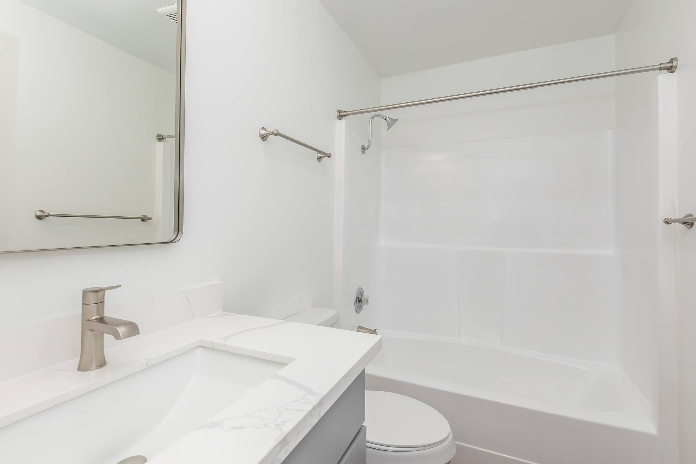 A clean and modern bathroom featuring a white tub and shower combination, a sleek sink with a minimalist faucet, and a large mirror above the sink. The walls are painted white, and there are metal towel bars installed. The overall look is bright and spacious.