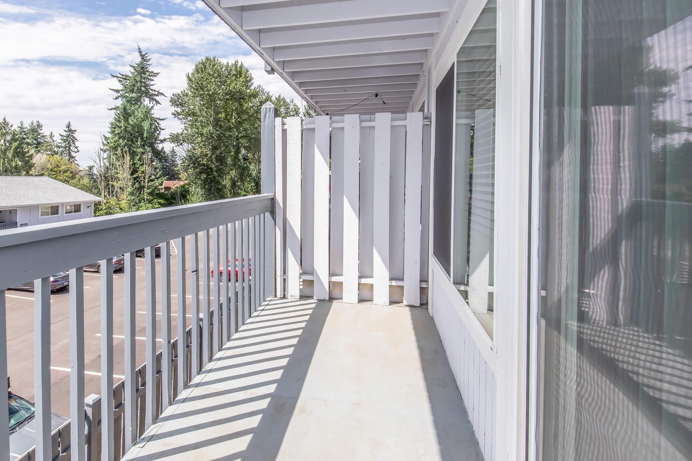 A white balcony with a railing, featuring a view of trees in the background and a parking area below. The floor is concrete, and there are shadows cast by the railing. The sky is partly cloudy, adding a serene atmosphere to the setting.
