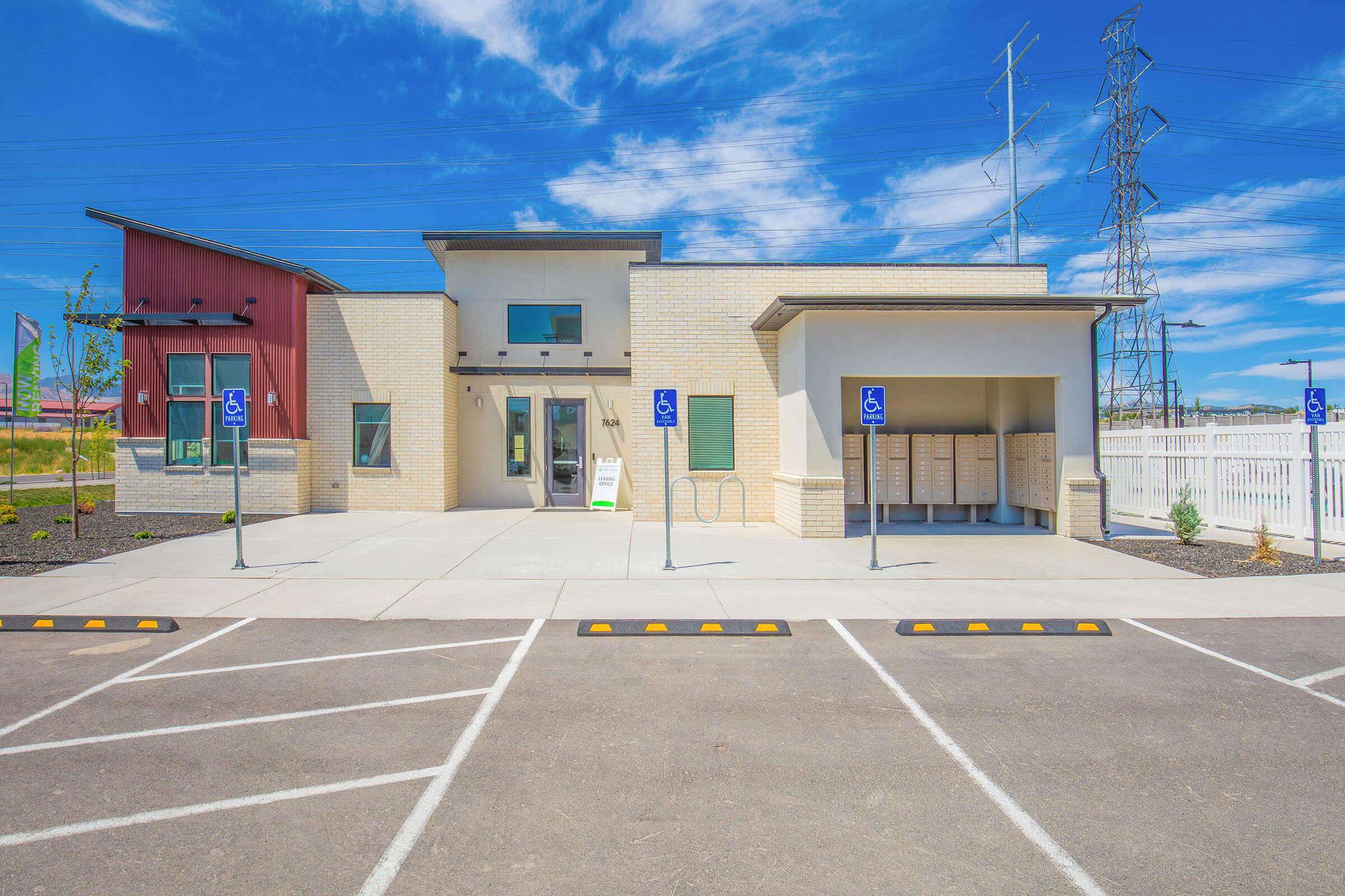 Modern building with a light brick exterior and red accents, featuring accessible parking spaces in front. The structure includes large windows and a flat roof. The landscape has minimal greenery, and there are bike racks near the entrance. Utility poles are visible in the background against a blue sky with scattered clouds.