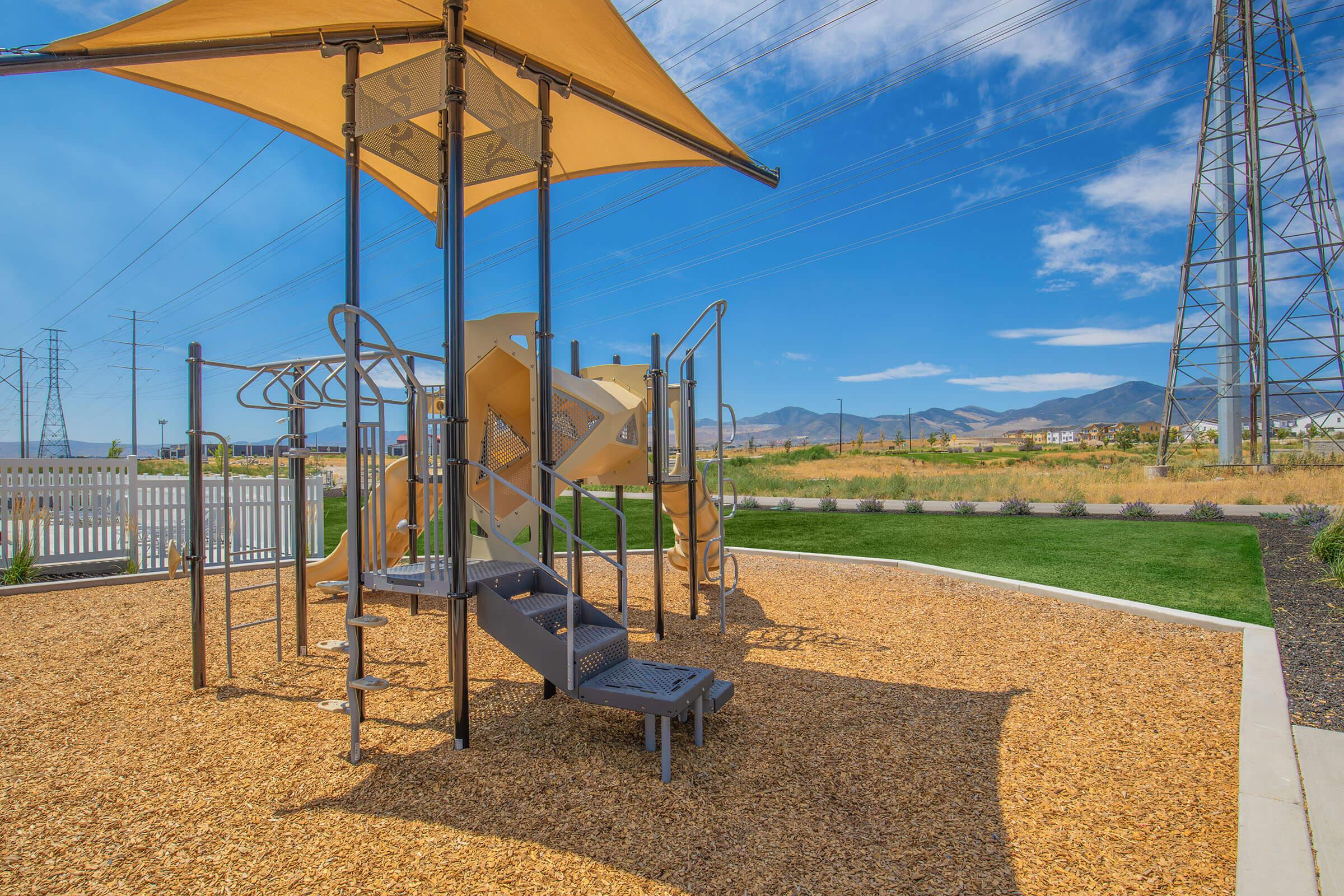 Playground equipment featuring slides, climbing structures, and a large shaded canopy, situated in a grassy area with wood chip flooring. In the background, utility poles and distant mountains are visible under a clear blue sky.