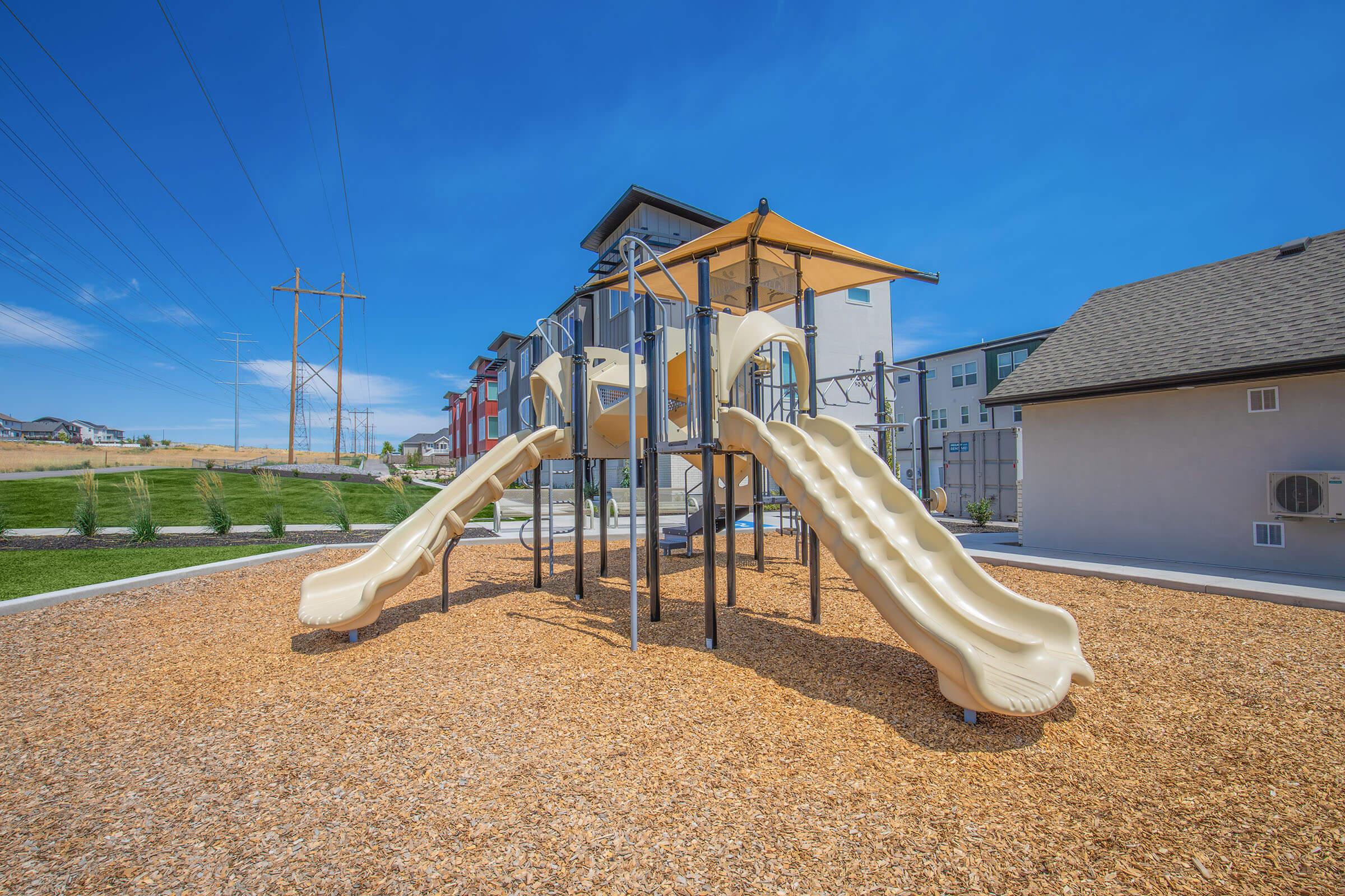 A playground featuring two beige slides, a climbing structure, and areas of wood chip flooring. The setting is bright with blue skies and power lines in the background, alongside residential buildings.