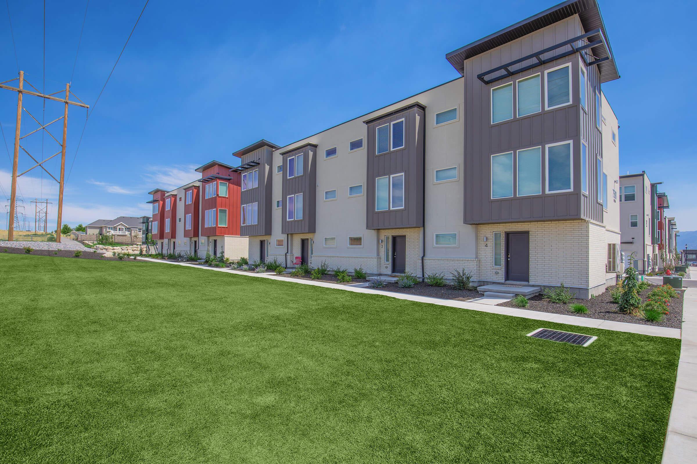 Modern multi-unit residential buildings with a variety of colors, set against a blue sky. The foreground features a lush green lawn, and utility poles can be seen in the background. The buildings have large windows and contemporary architectural designs.