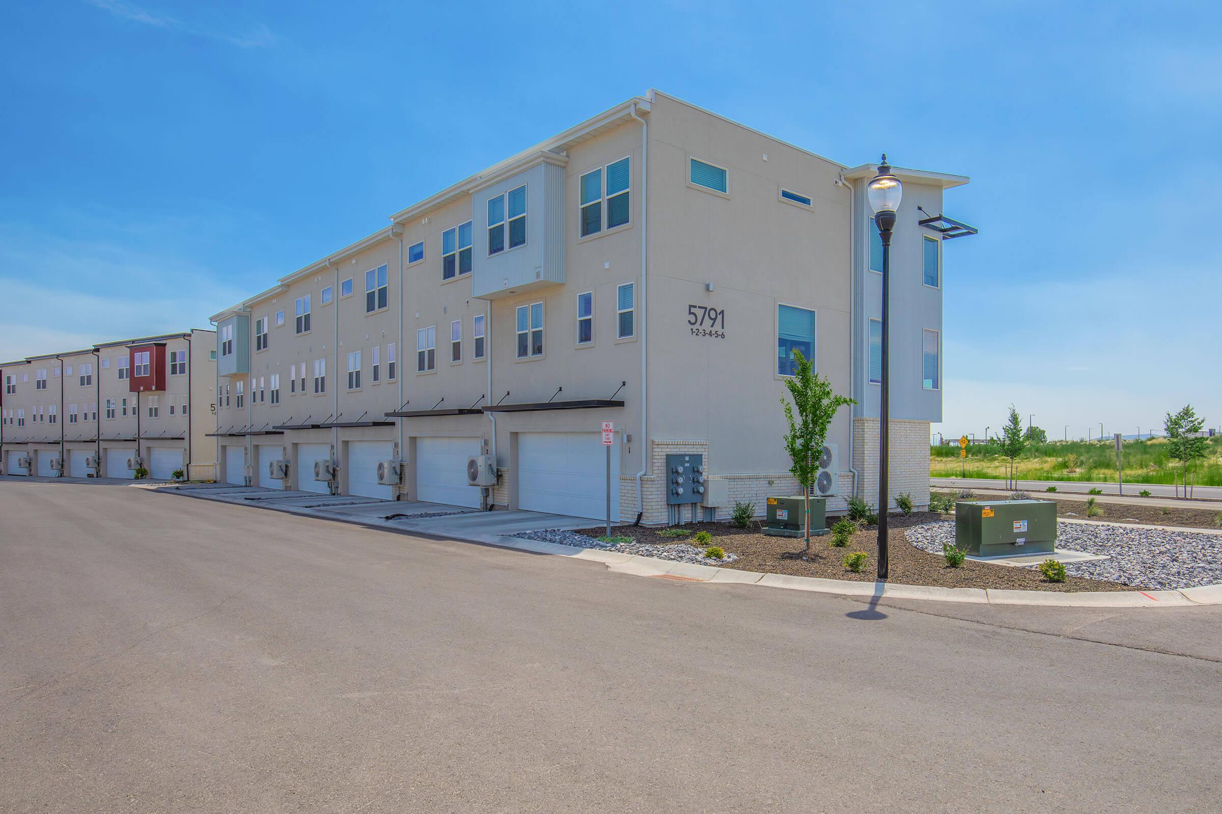 A modern, multi-story residential building with several garage doors, located at 5791. The architecture features large windows and clean lines, set against a clear blue sky. The surrounding area is landscaped with small shrubs and grass, with utility boxes visible on the ground.
