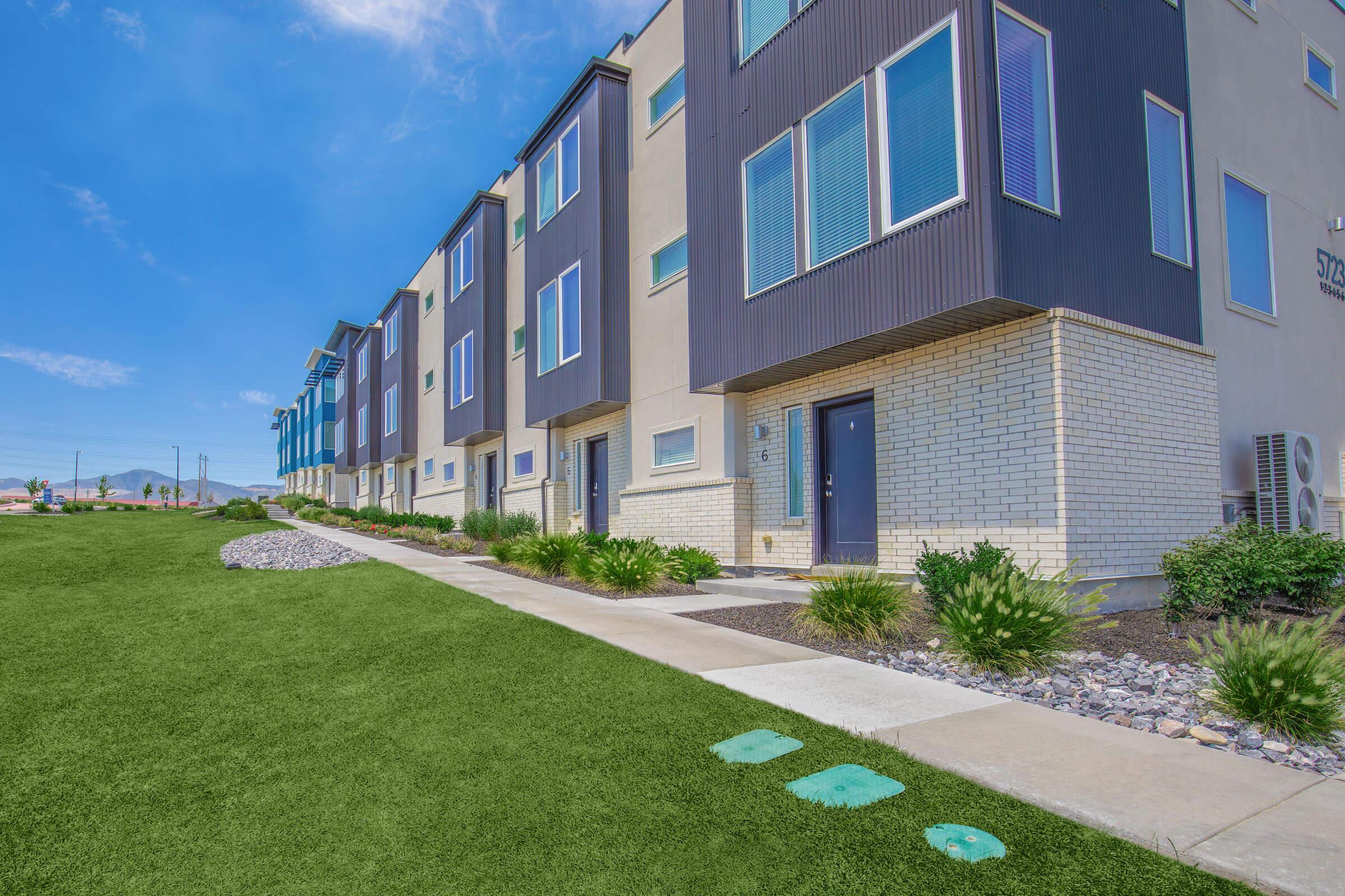A row of modern townhouses with a mix of beige and dark exterior, featuring large windows and small front gardens. A manicured lawn runs alongside a concrete walkway, leading to the entrances. The sky is clear and blue, with a few clouds visible in the background.