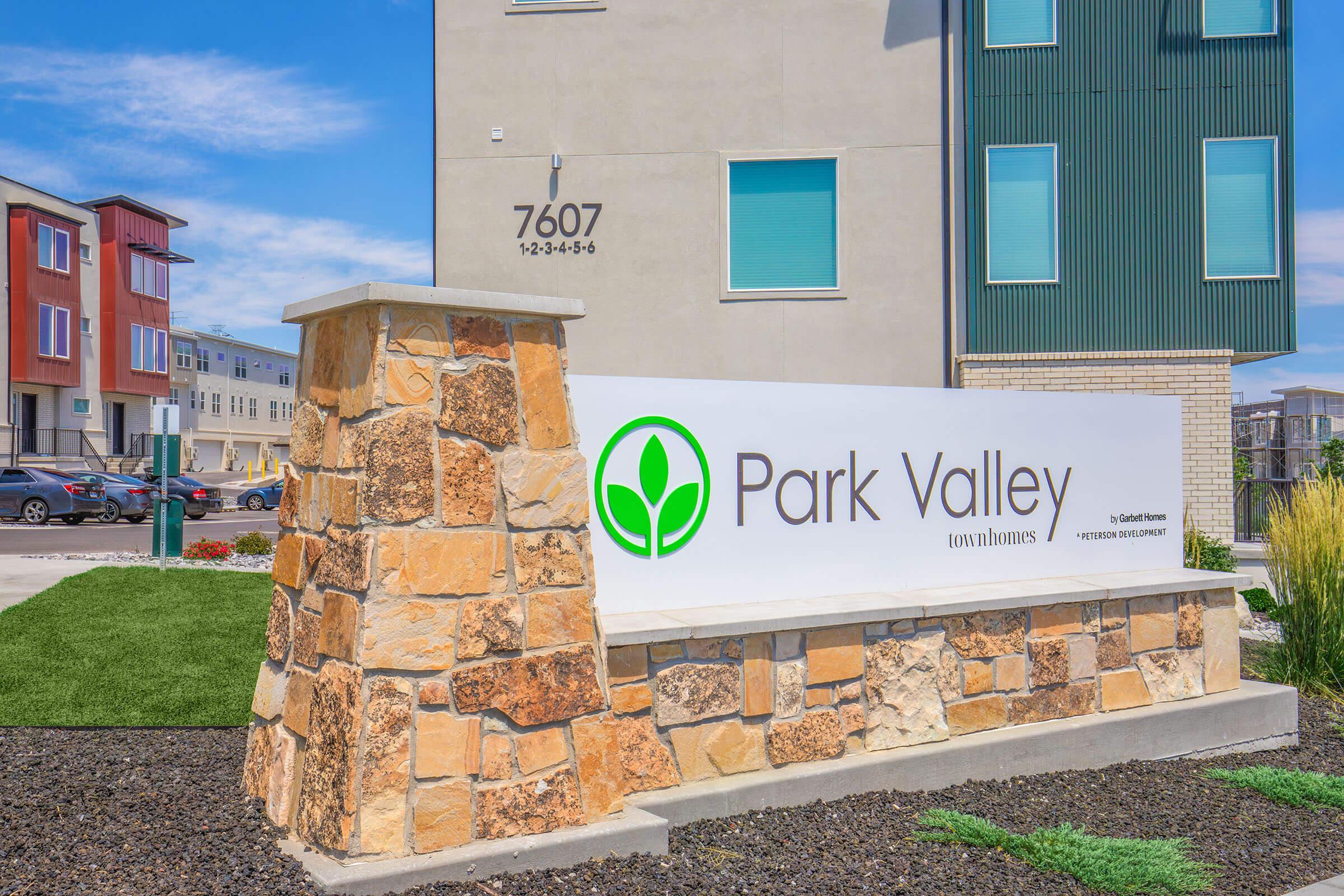 Sign for Park Valley townhomes featuring a green logo with a leaf design. The sign is made of stone and is prominently displayed outside a modern multi-story building. The building address, 7607, is visible above the sign. Landscaping with grass and decorative stones surrounds the area.