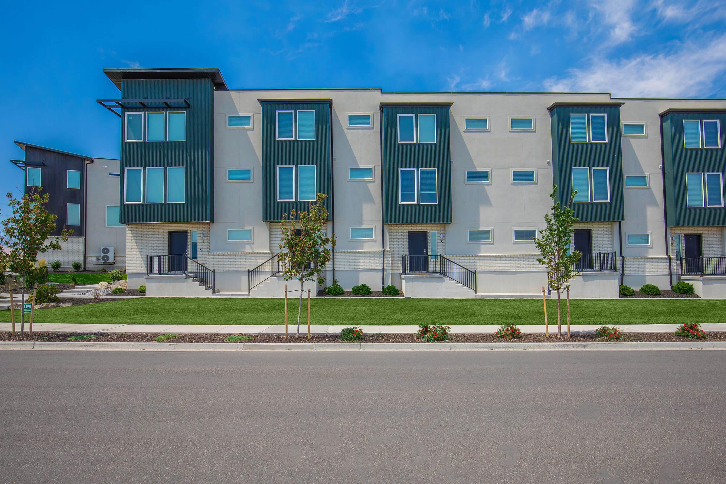 Modern three-story residential building with a minimalist design, featuring a combination of light and dark exterior colors. Large windows line the front, with a well-maintained lawn and small shrubs in front. A clear blue sky is visible above, enhancing the contemporary aesthetic of the property.