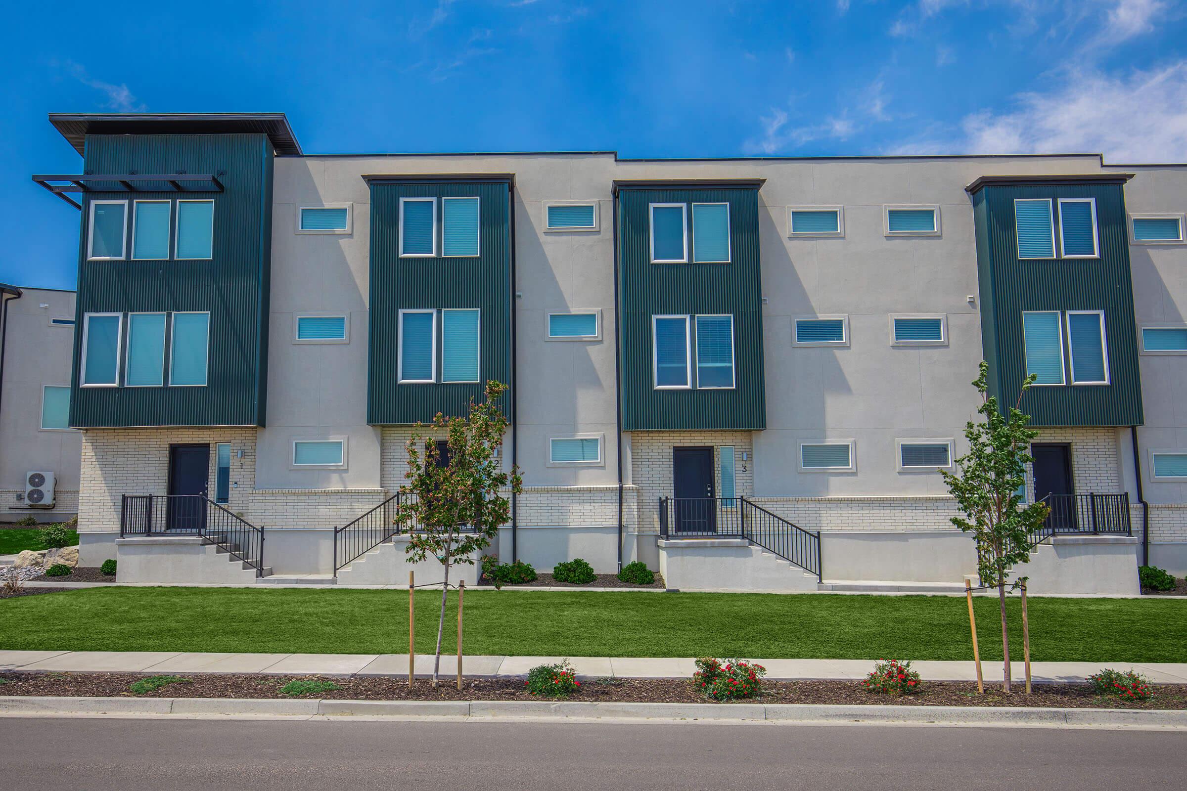 A modern two-story residential building featuring a row of three units with large windows and a clean, minimalist design. The exterior is a mix of light-colored stucco and dark paneling. In front, there is a well-maintained lawn with small bushes and flowers, creating a welcoming atmosphere.