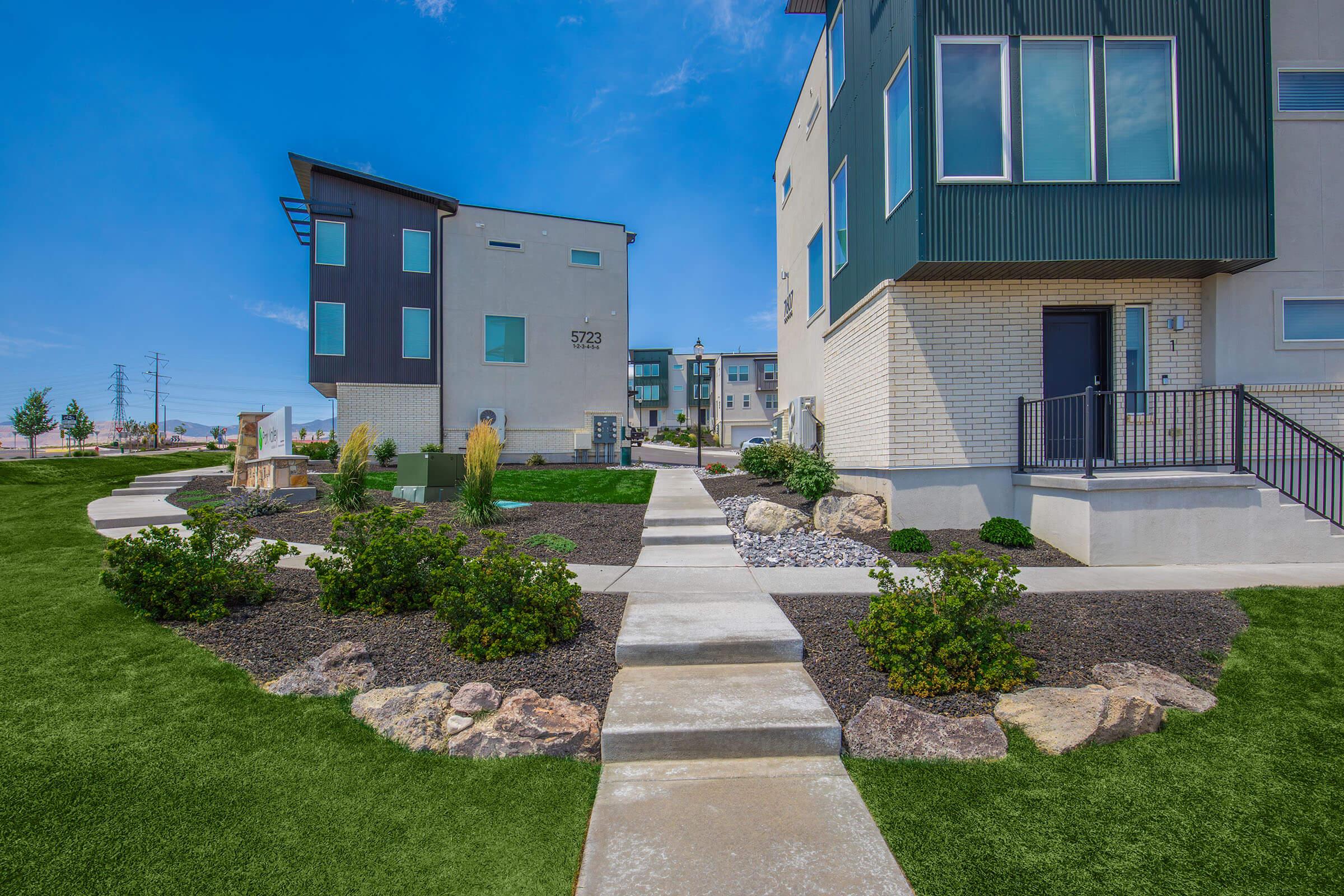 A modern residential neighborhood featuring two contemporary-style homes with contrasting exteriors. A paved walkway leads through a landscaped yard with rocks and greenery. The sky is clear and blue, creating a bright atmosphere. Power lines are visible in the background, indicating a suburban setting.