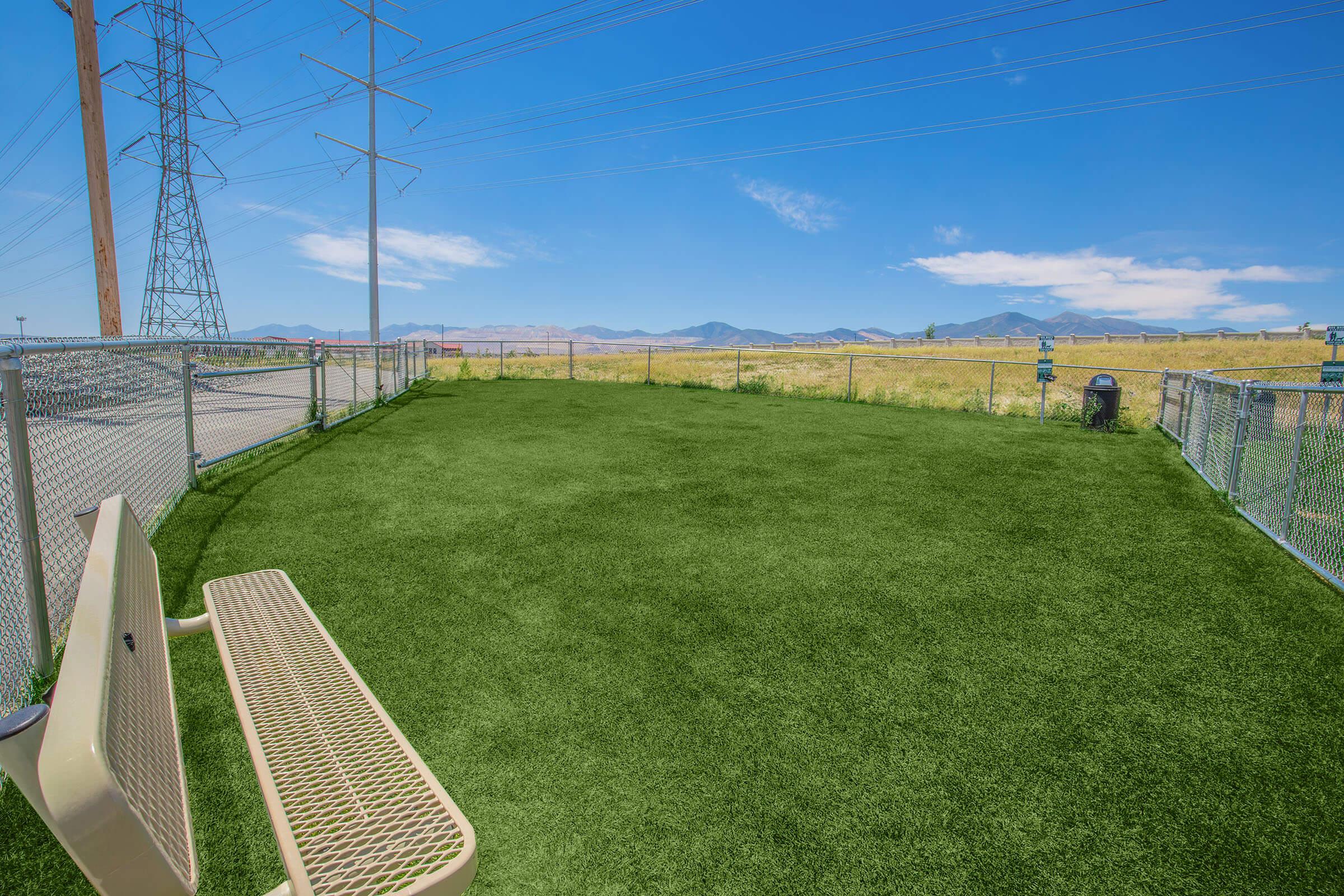 A spacious dog park with artificial grass, featuring a white bench near a chain-link fence. Power lines stretch across a clear blue sky, and mountains are visible in the distance. The park appears well-maintained and is surrounded by open fields.