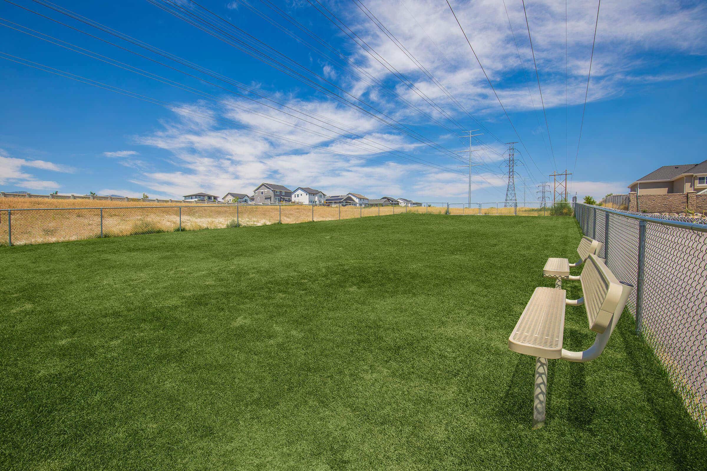 A green grassy area enclosed by a fence, featuring two benches. In the background, there are houses and power lines under a bright blue sky with scattered clouds.