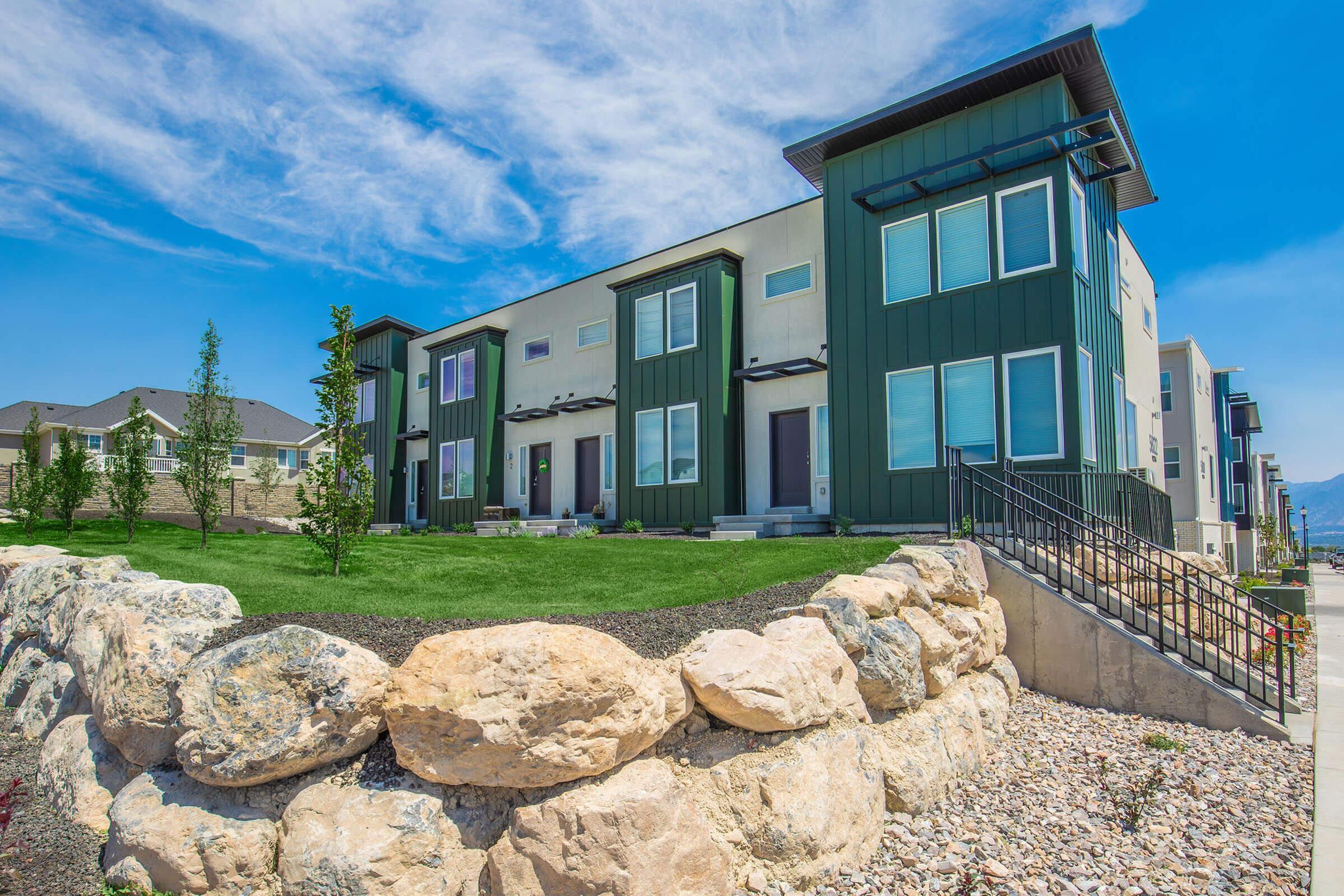 Modern green and beige residential buildings with multiple units, featuring large windows and balconies. The foreground includes a landscaped area with a stone wall and grass. The background shows a clear blue sky with some clouds.