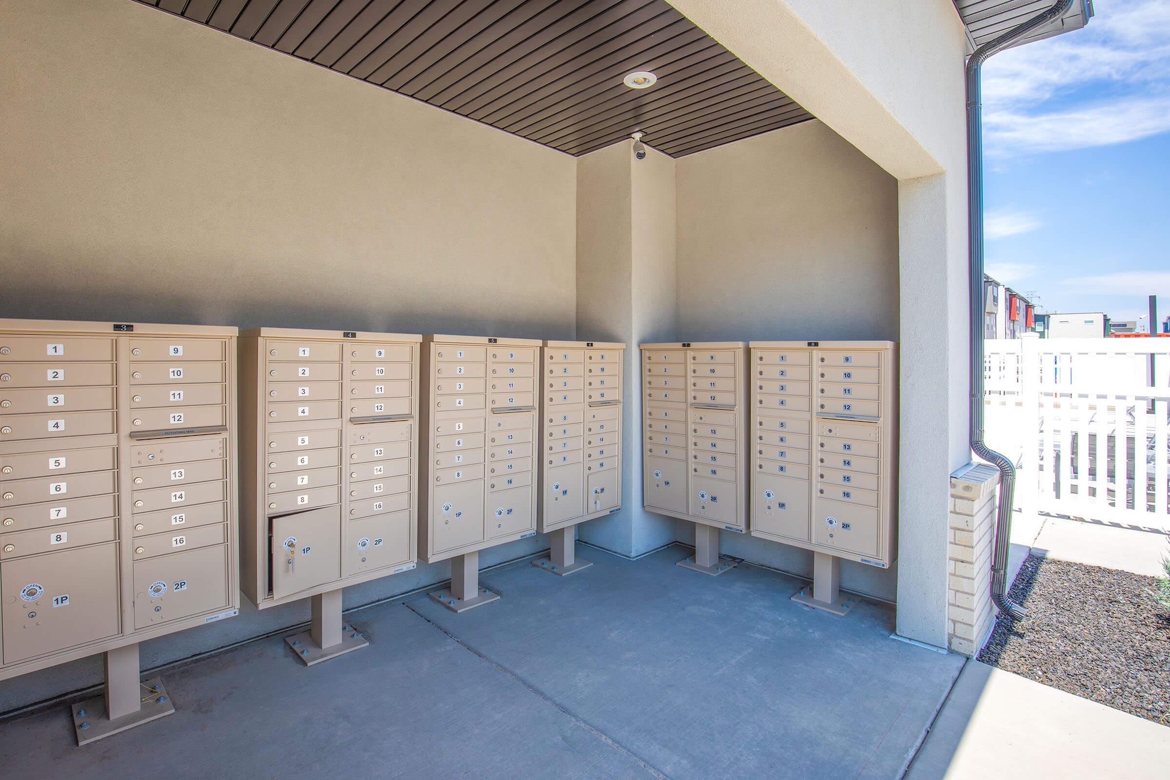 A row of beige mailboxes installed outside a residential building, set against a light gray wall. The mailboxes are neatly arranged and mounted on metal stands, with a clear blue sky in the background. The area is well-lit and clean, suggesting a modern residential community.