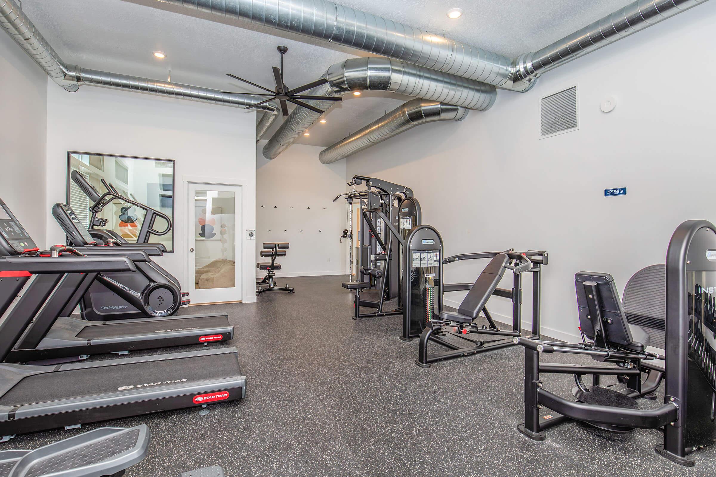 A modern home gym featuring multiple exercise machines including treadmills, strength training equipment, and a variety of fitness tools. The space has rubber flooring, bright lighting, and an industrial ventilation system with exposed ducts. A mirror is visible on one wall, enhancing the workout experience.