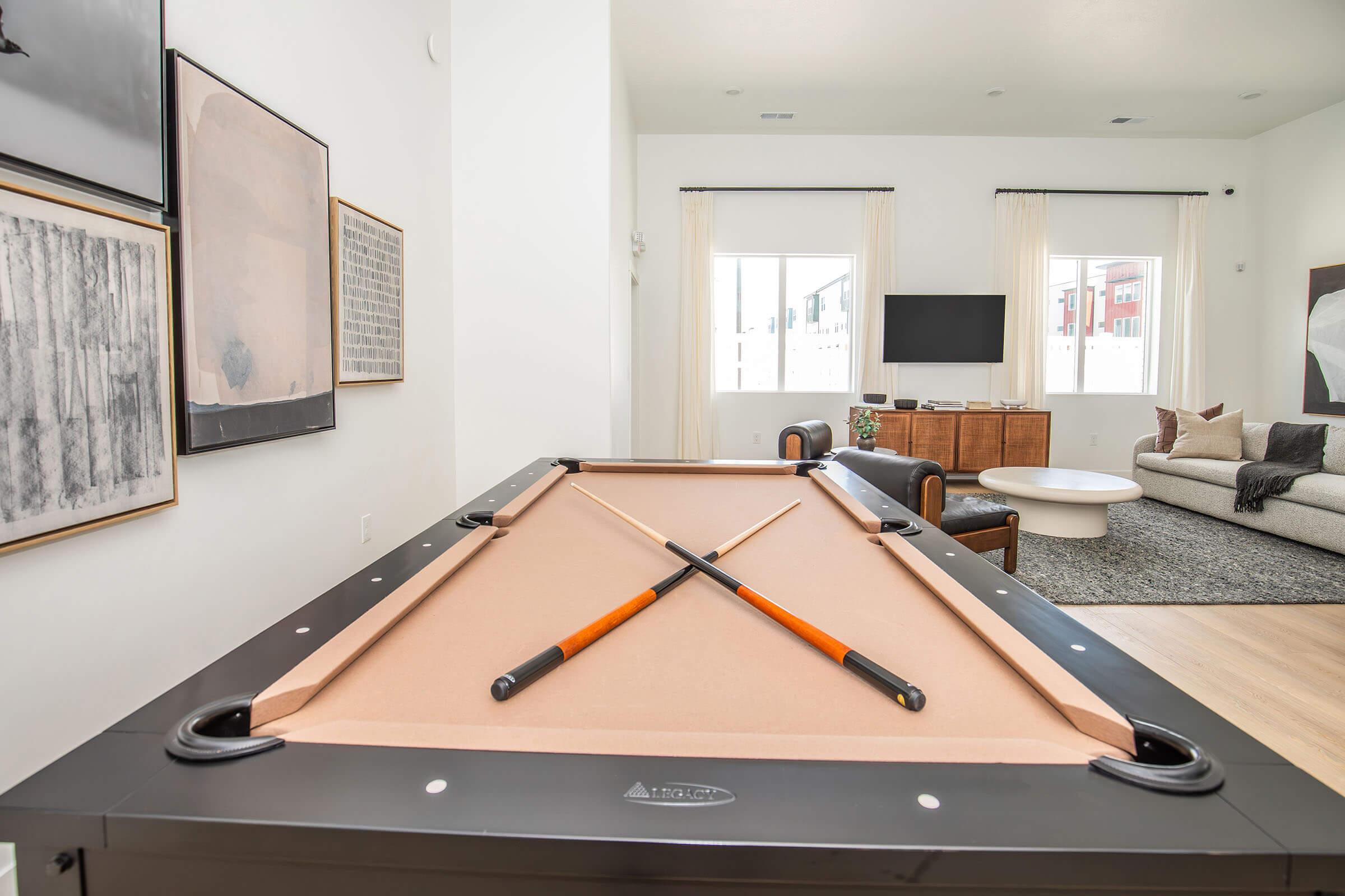A spacious interior featuring a billiard table with crossed cue sticks on top. In the background, there are windows with natural light, a modern TV mounted on the wall, and stylish furniture including a couch and a coffee table, enhancing the contemporary decor of the room.