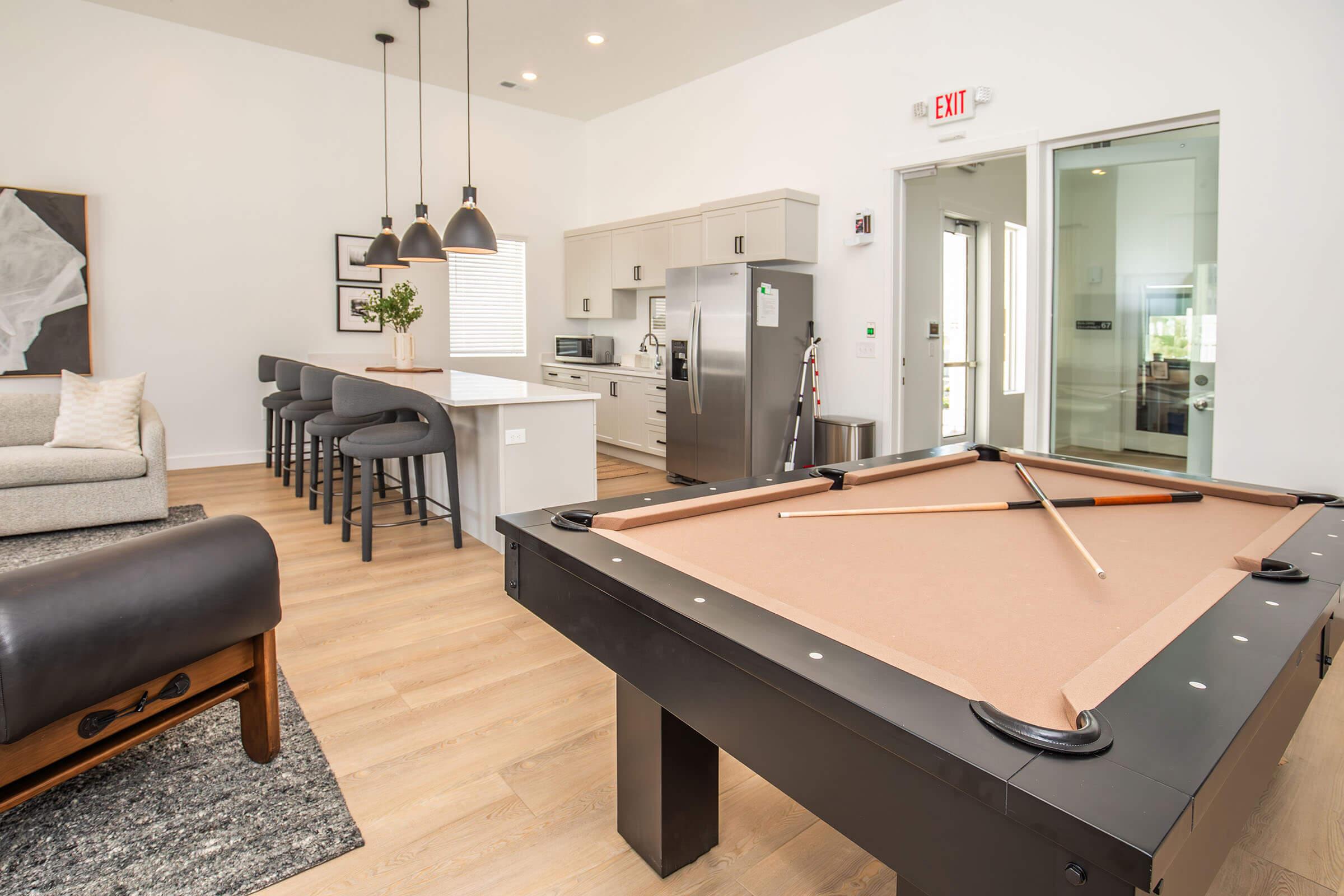 A modern living space featuring a pool table in the foreground, with a stylish sofa and bar stools in the background. The open kitchen includes stainless steel appliances and contemporary decor, with large windows allowing natural light to fill the room.