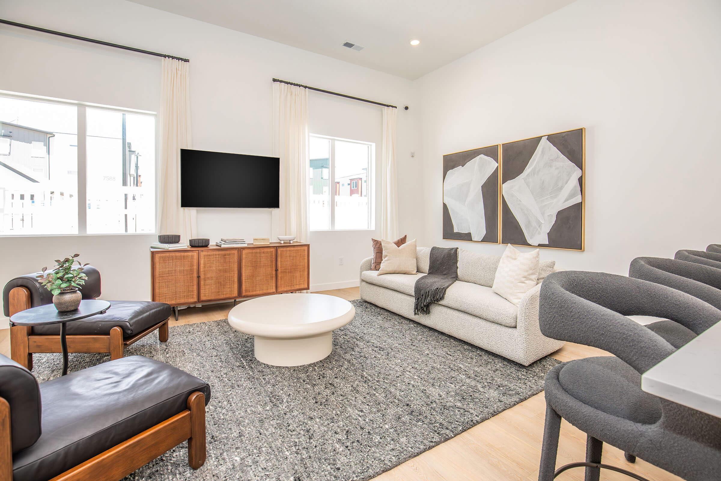 A modern living room featuring a white sofa with decorative pillows, a round coffee table, and two black armchairs. The room has a textured gray rug, large windows, and minimalistic artwork on the walls. There’s also a television mounted on the wall and a wooden side cabinet.