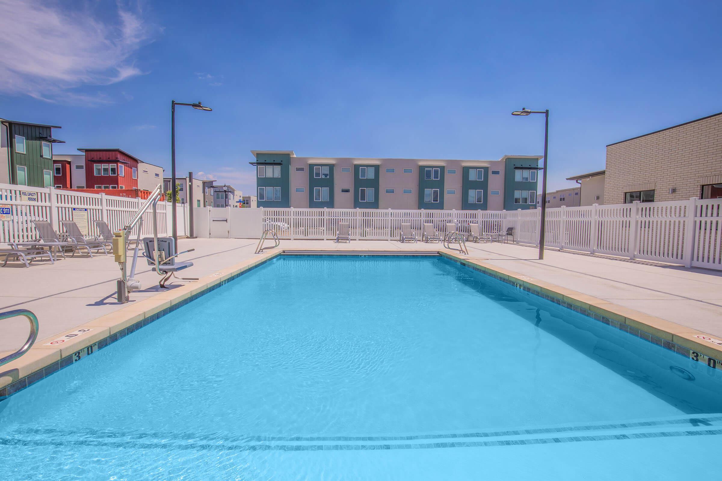A bright, inviting swimming pool surrounded by a white fence, with lounge chairs arranged neatly on the pool deck. In the background, colorful apartment buildings are visible under a clear blue sky, creating a vibrant and modern atmosphere.