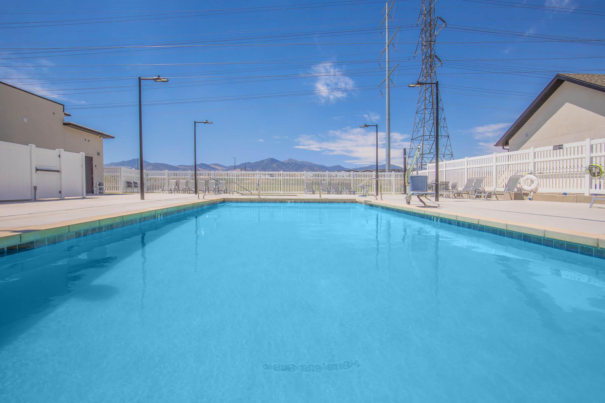 A clear blue swimming pool with a surrounding deck, framed by white fences and light poles. In the background, mountains are visible under a bright blue sky with a few clouds. Power lines run across the scene, adding to the outdoor atmosphere.