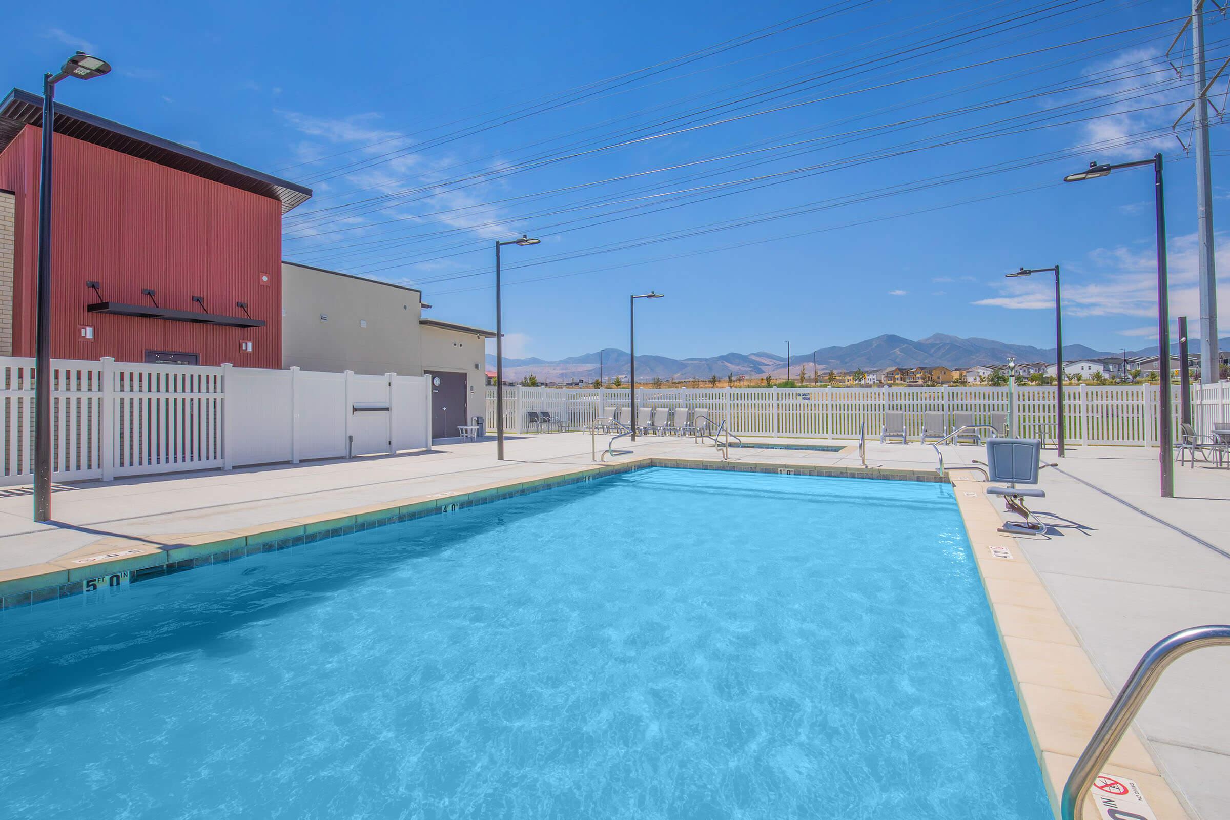 A clear swimming pool with light blue water is surrounded by a concrete deck. There are lounge chairs and tall light poles nearby, with a white fence in the background. The scene is set under a bright blue sky with a few clouds and mountains visible in the distance. Power lines run across the sky.