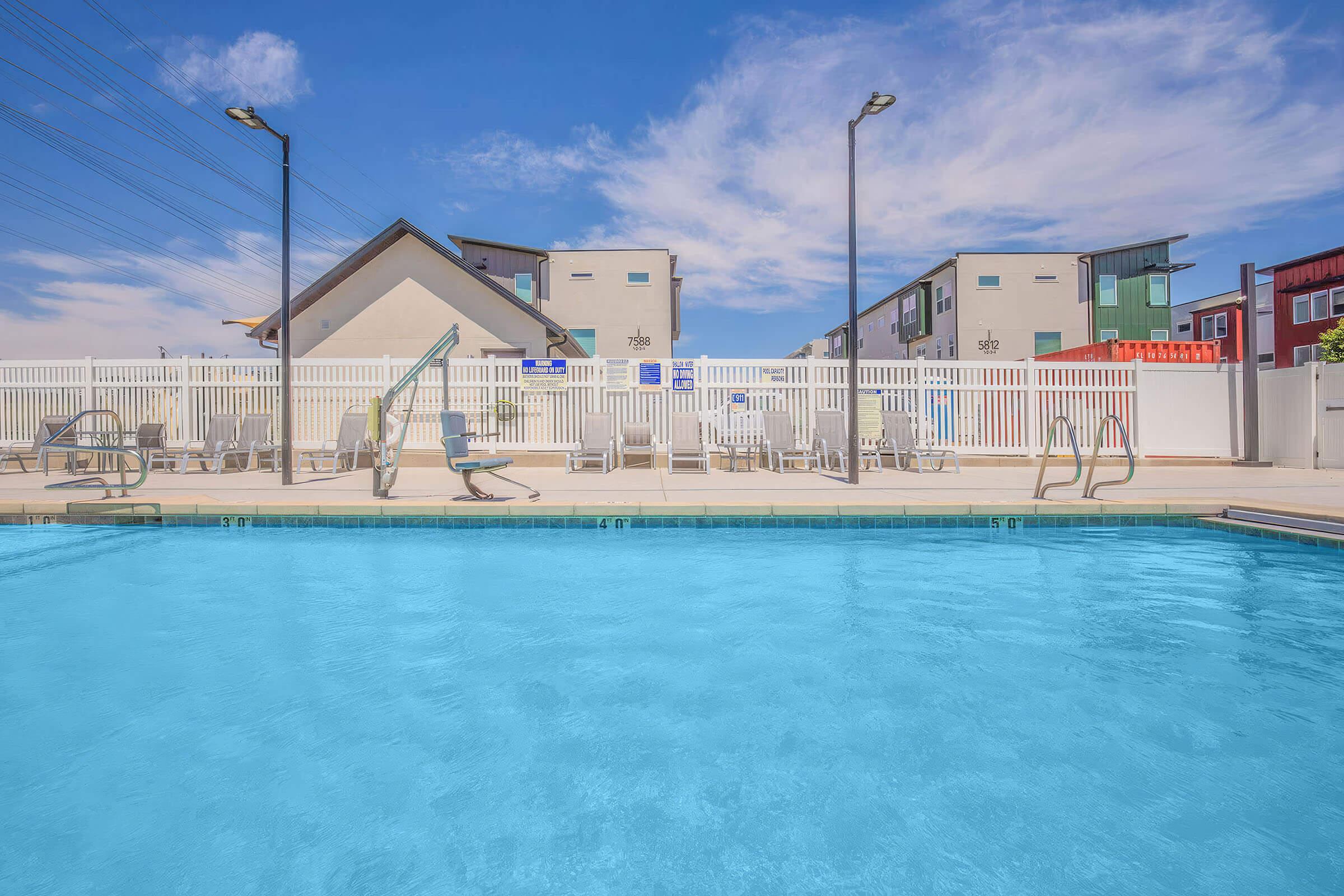 A clear blue swimming pool surrounded by lounge chairs and a white fence. In the background, modern buildings with a mix of colors are visible under a bright blue sky with scattered clouds. Two poolside ladders are positioned at opposite ends of the pool.
