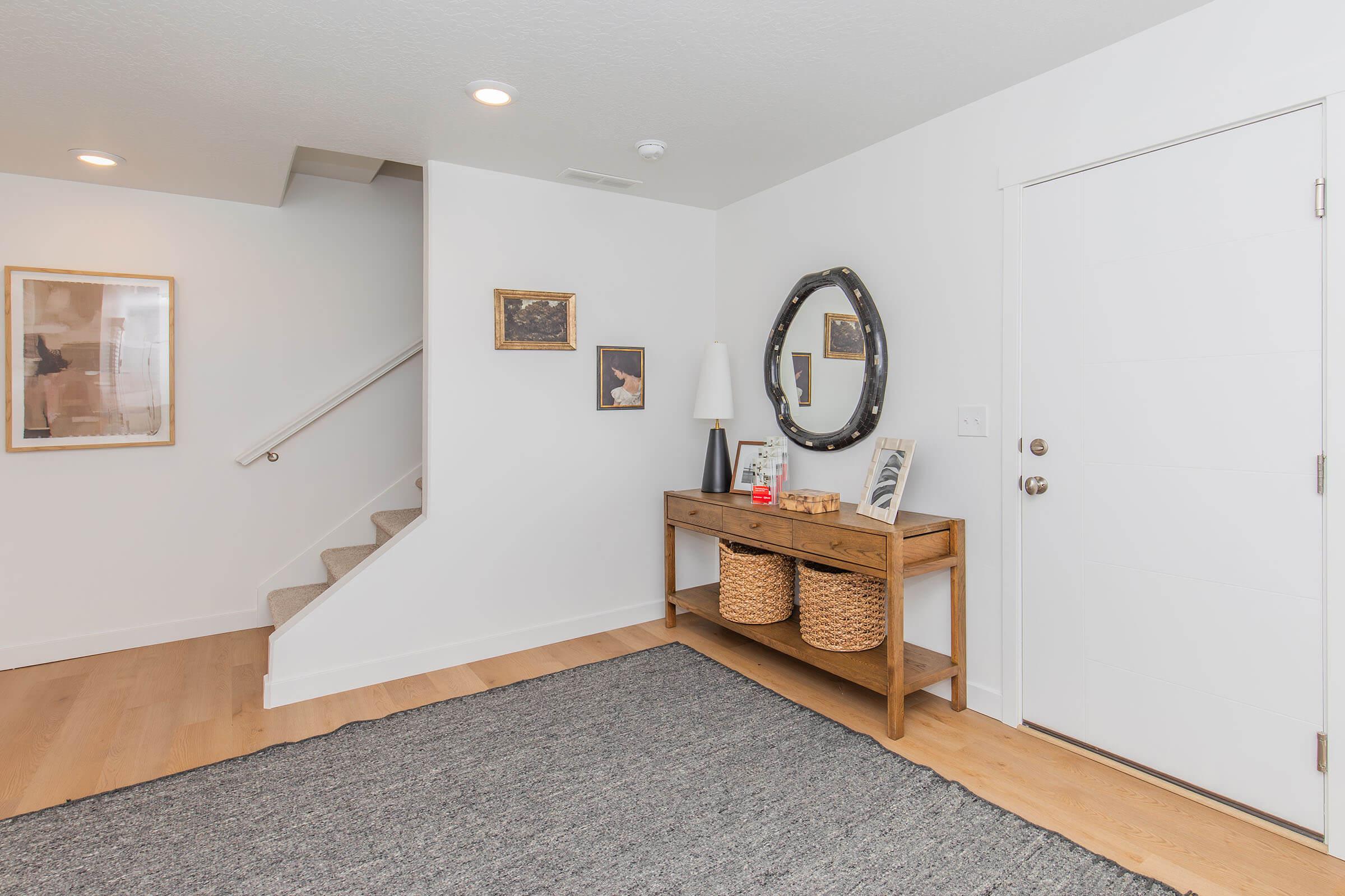 A modern entryway featuring a wooden console table with two woven baskets underneath. Above the table are framed artworks and a round mirror. The wall is white, and the space includes a staircase in the background, illuminated by recessed lighting, with a textured gray rug on the floor.