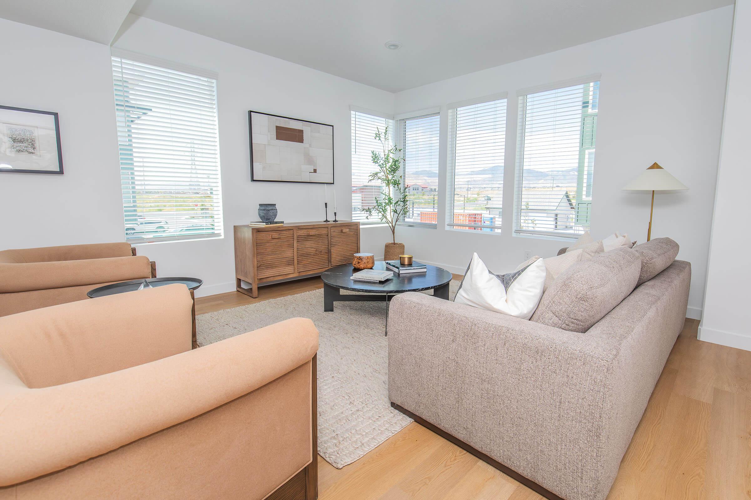 A modern living room with large windows allowing natural light, featuring two beige armchairs, a cozy light gray sofa with decorative pillows, a round coffee table in the center, and a wooden sideboard against the wall. A houseplant adds a touch of greenery to the space, creating a warm and inviting atmosphere.