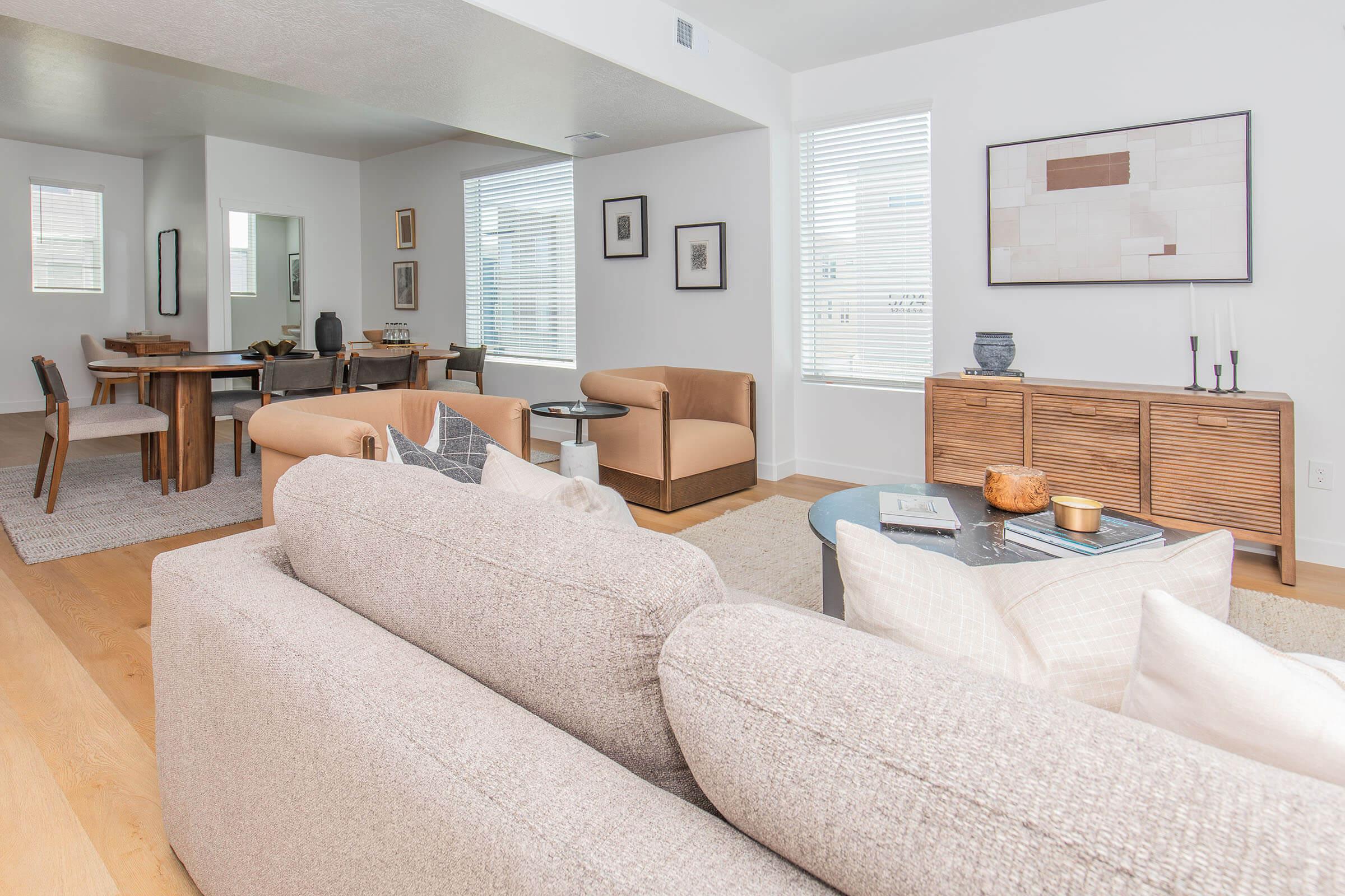 A modern, stylish living room featuring a light-colored sofa, two tan armchairs, and a coffee table with decorative items. The space has wooden flooring and minimalist decor, including artwork on the walls and a dining area visible in the background with a table and chairs. Large windows allow natural light to fill the room.