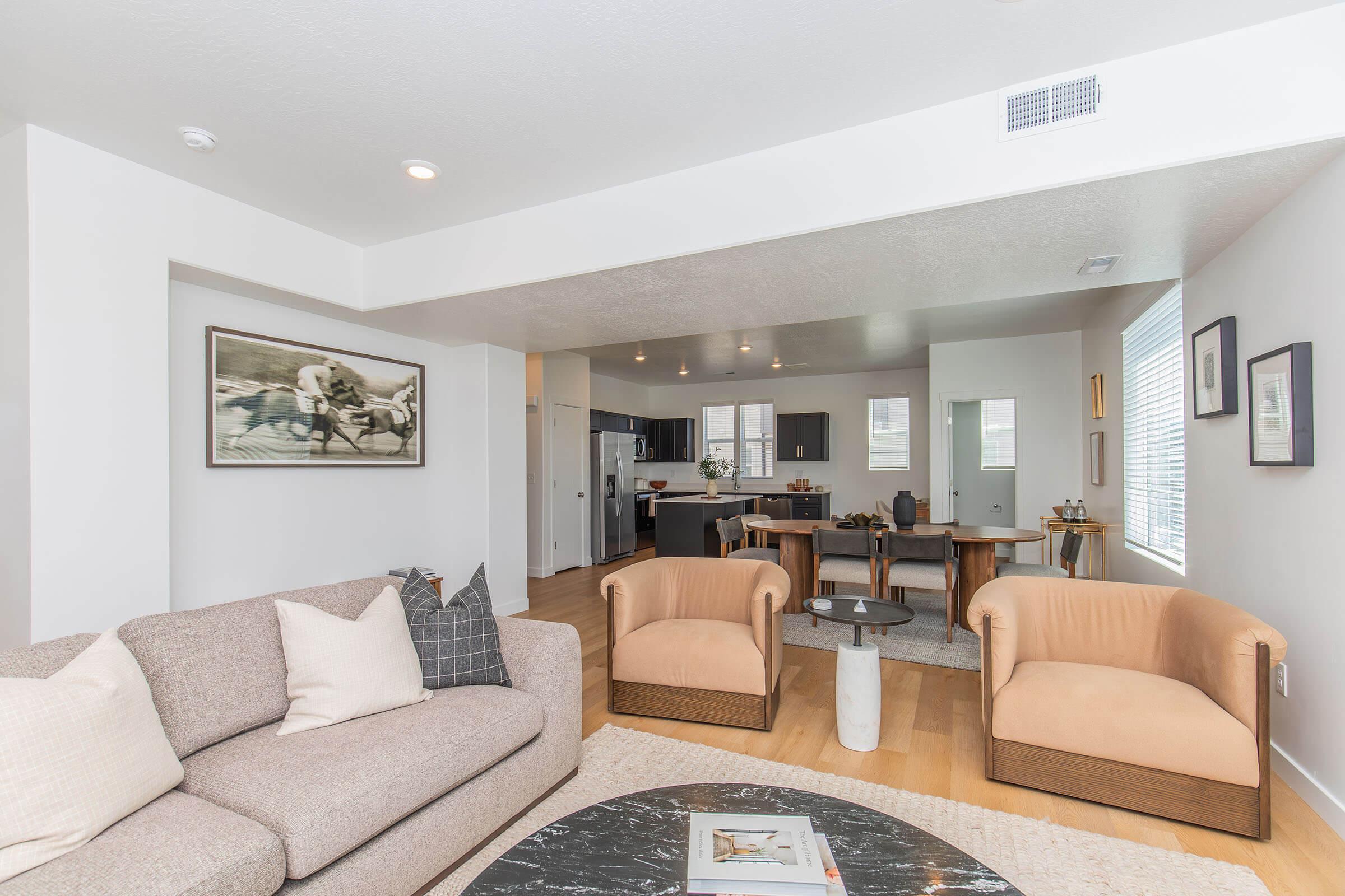 A modern, stylish living room featuring a beige sectional sofa, two brown armchairs, and a round black coffee table. The walls are painted white, with a framed horse photograph and decorative accents. A dining area with wooden table and chairs is visible in the background, alongside a kitchen with dark cabinetry.